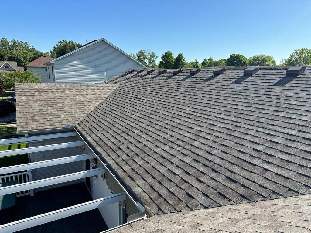 Overhead view of a roof with dark gray shingles and a brick wall. Sunlight, blue sky.
