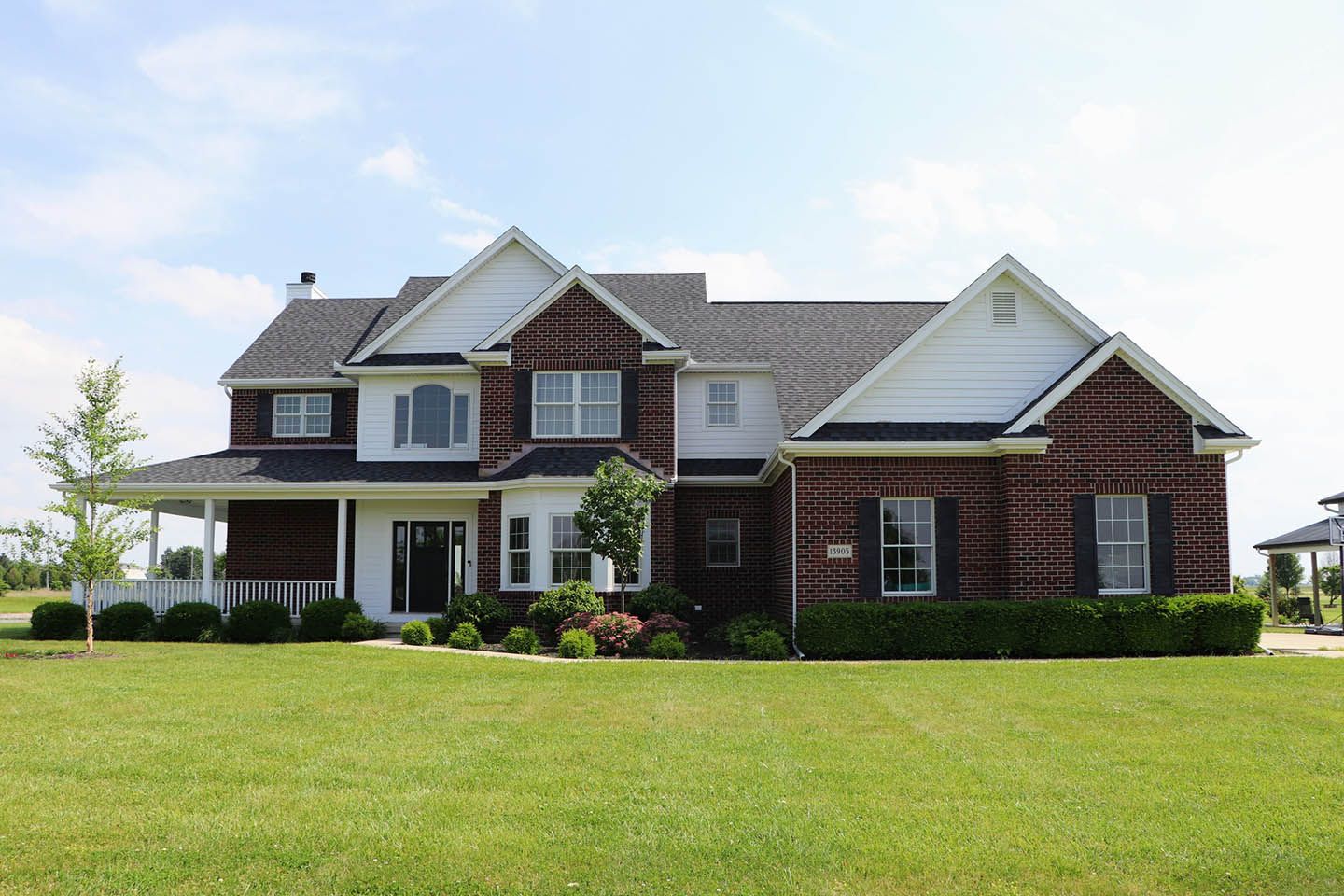 Two-story brick house with white trim, black shutters, and a large front lawn under a blue sky.