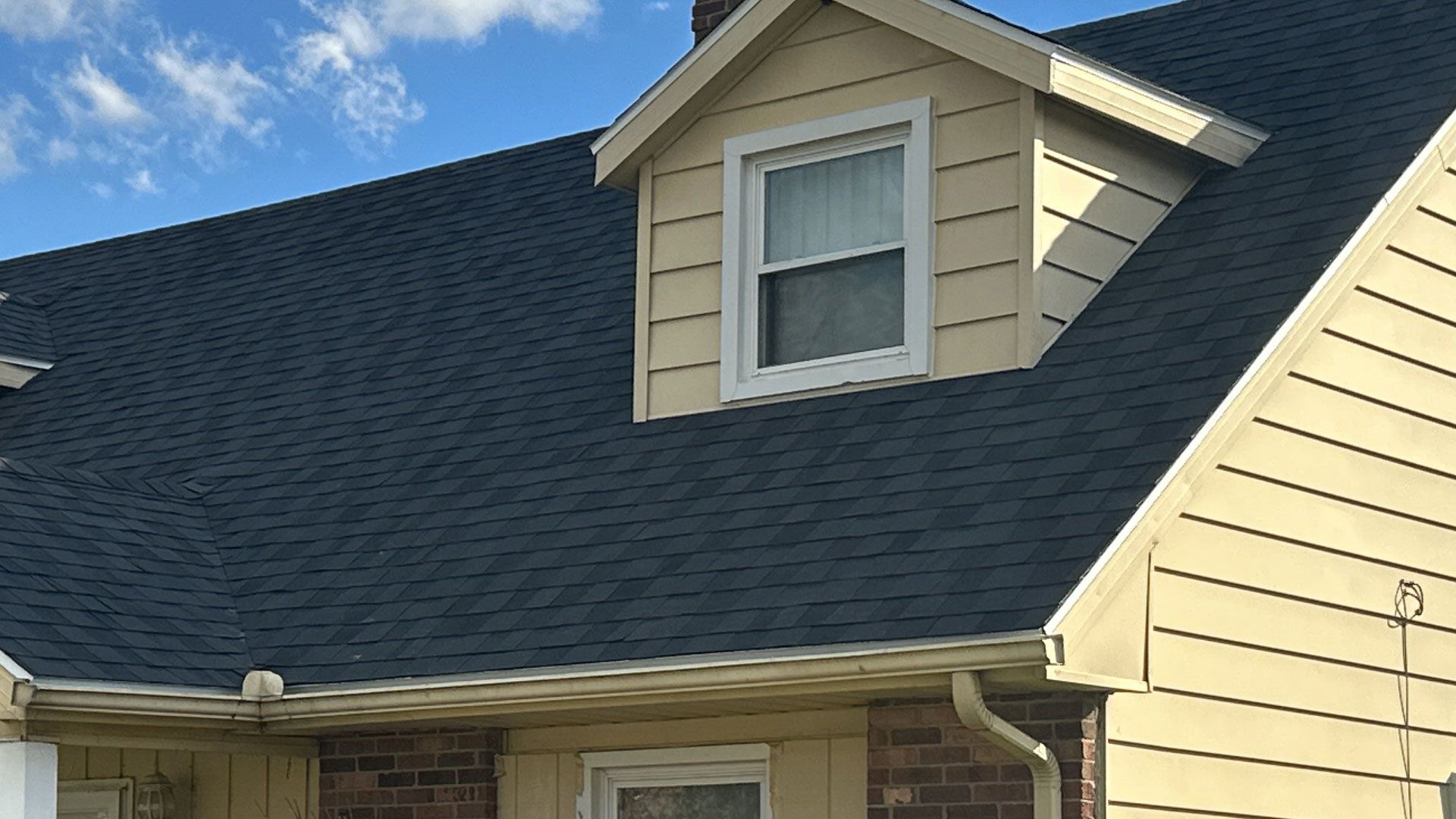 Dark blue shingle roof with a light yellow dormer and a white-framed window against a blue sky.