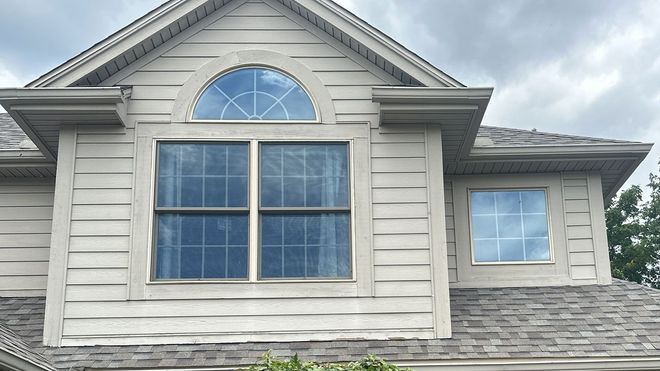 Beige house with multiple windows and a curved window at the top. Gray roof and cloudy sky.