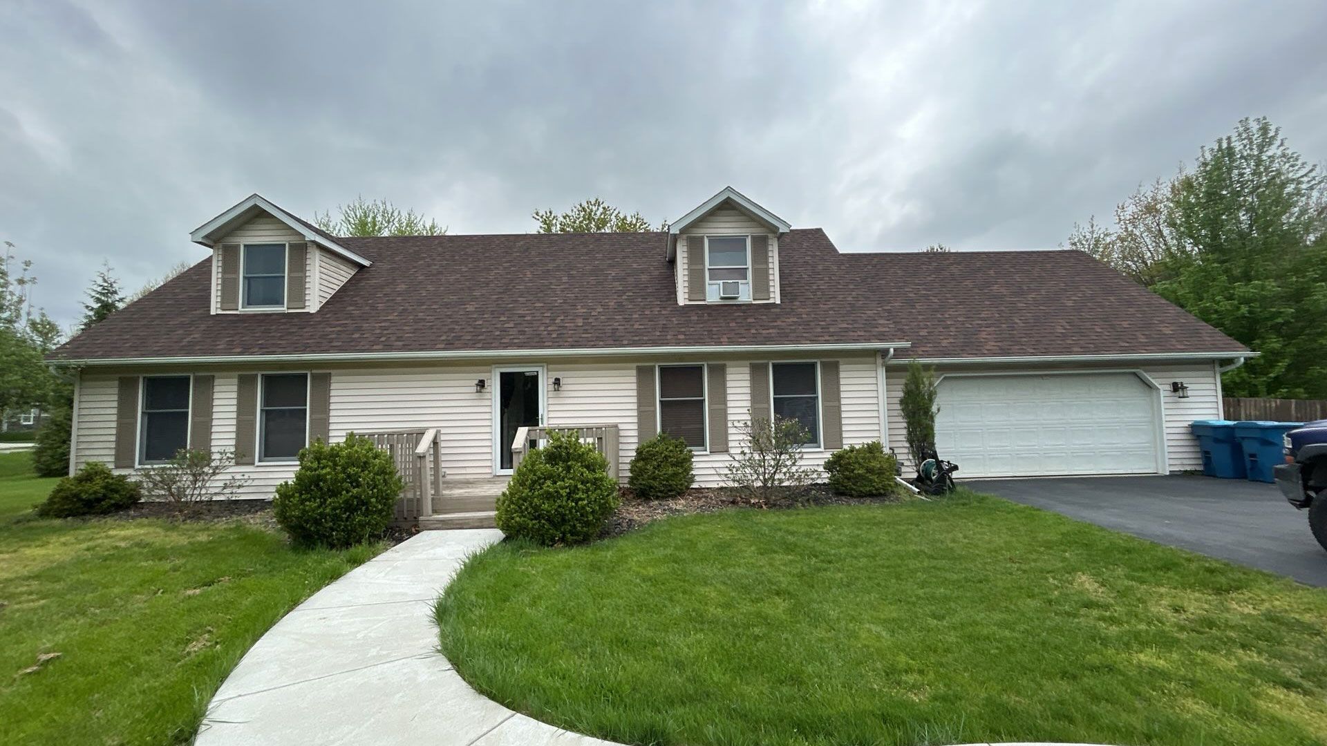 Ranch-style house with dormers, light siding, brown roof, and a white garage door. A curved sidewalk leads to the front door.