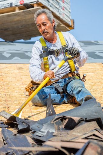 Roofer kneeling, removing shingles with a pry bar, wearing safety harness. OSB roof, blue jeans.