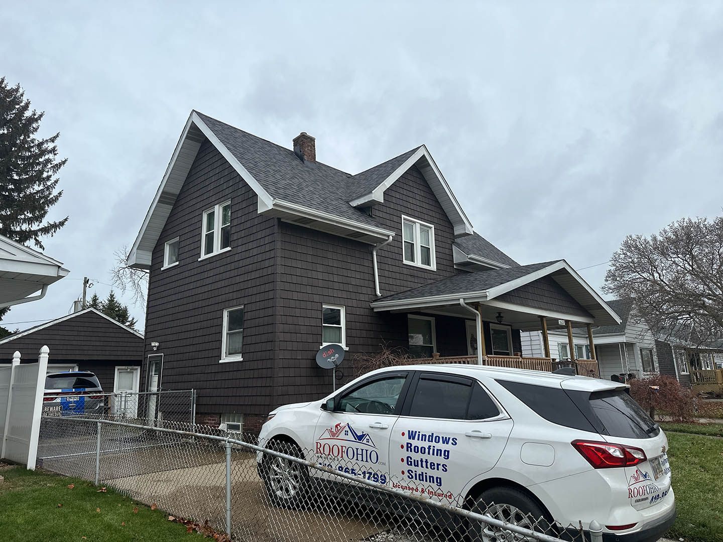 Brown house with new roof, white trim. White SUV with business logo in front of the house. Overcast sky.