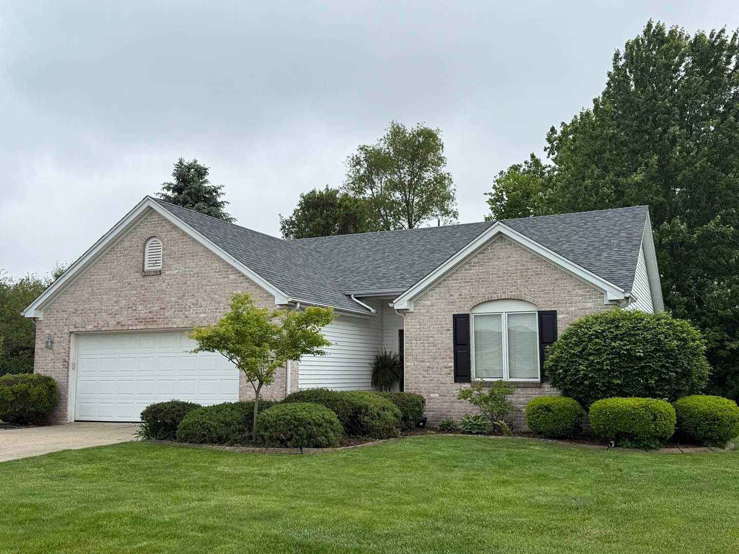 Brick house with gray roof, white garage door, black shutters, and green bushes. Overcast sky.