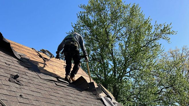 Roofer in safety harness working on a partially demolished roof under a blue sky, tree in background.