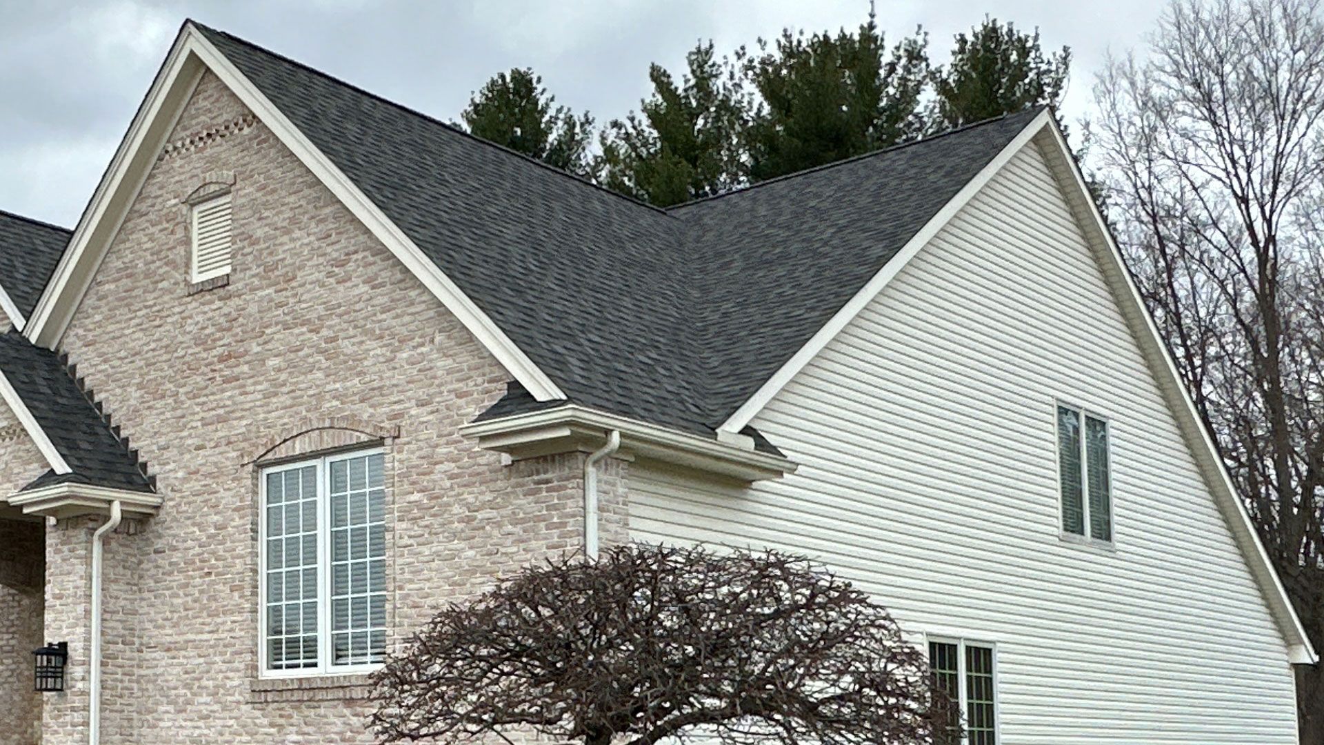 House with gray shingle roof, beige brick and white siding, white trim, cloudy sky.