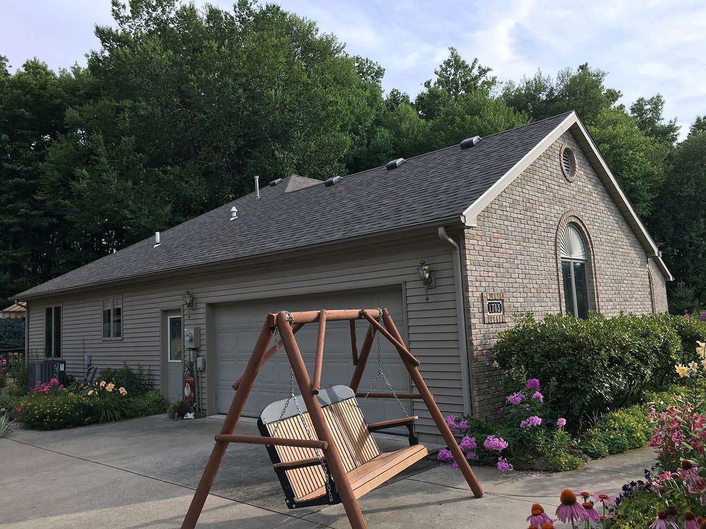 House with gray roof, tan siding, and a wooden swing in front. Lush green trees and flowers surround it.