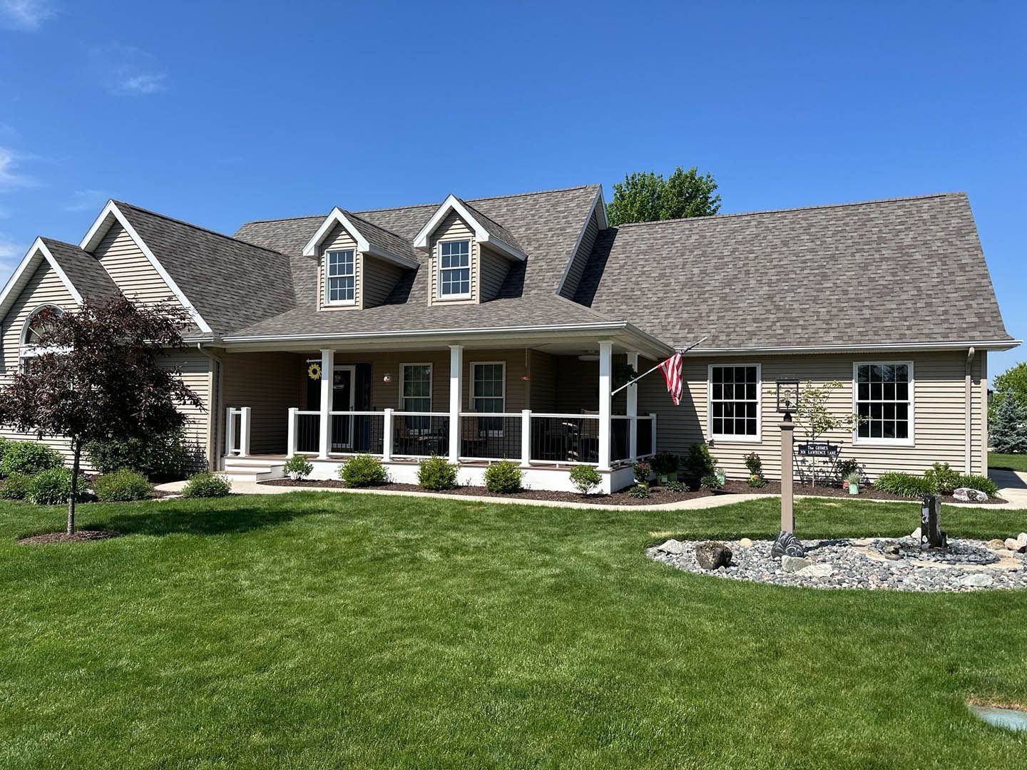 Ranch-style house with gray roof, porch, and dormers. Green lawn, blue sky, flag.
