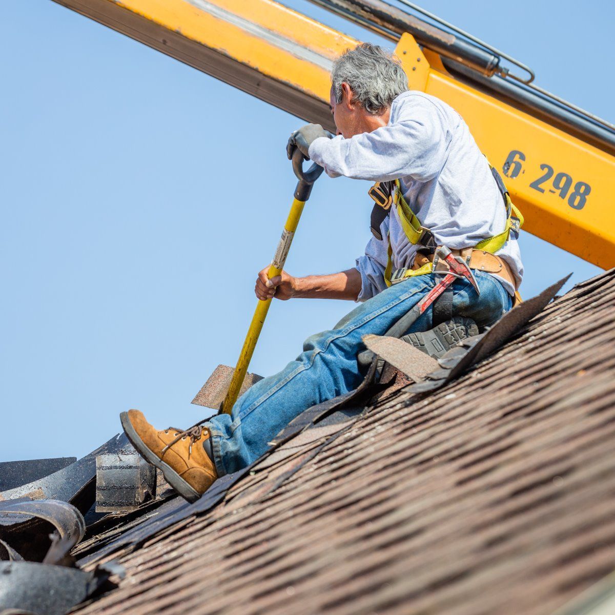 Roofer using a shovel to remove shingles from a roof, wearing a safety harness, yellow lift behind.