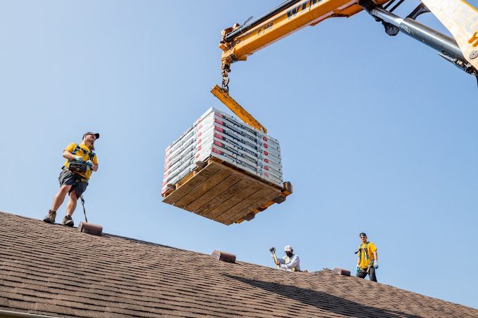 Crane lifting shingles to a roof; three workers watch, one operating a remote control.