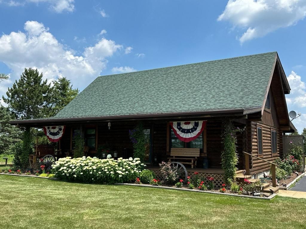 Log cabin with green roof, porch decorated with patriotic bunting, surrounded by flowers and green lawn under a blue sky.