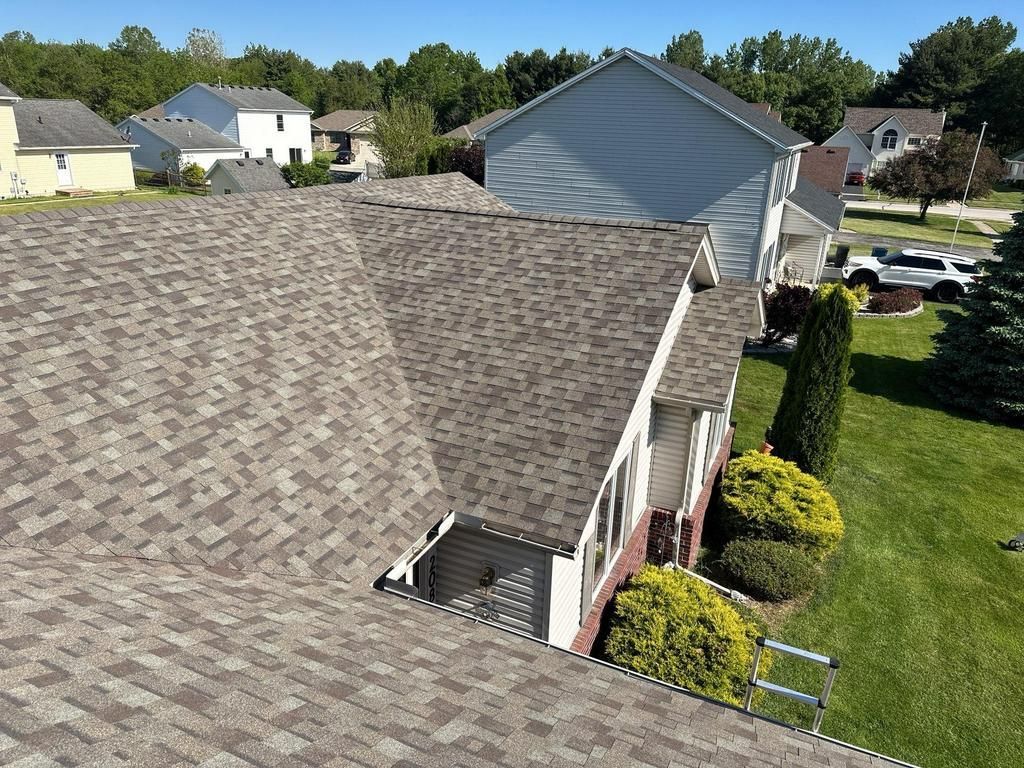 Newly shingled roof of a house; brown and gray shingles. Sunlight, blue sky, and green grass.