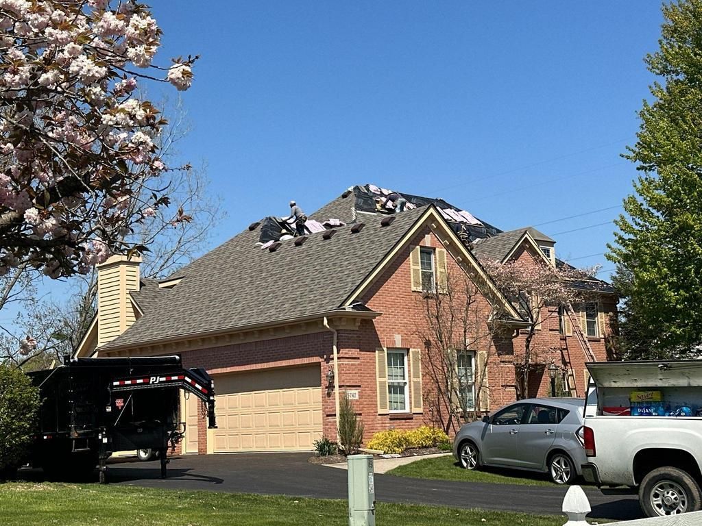 Workers on a roof replacing shingles, brick house with a garage, vehicles in the driveway.