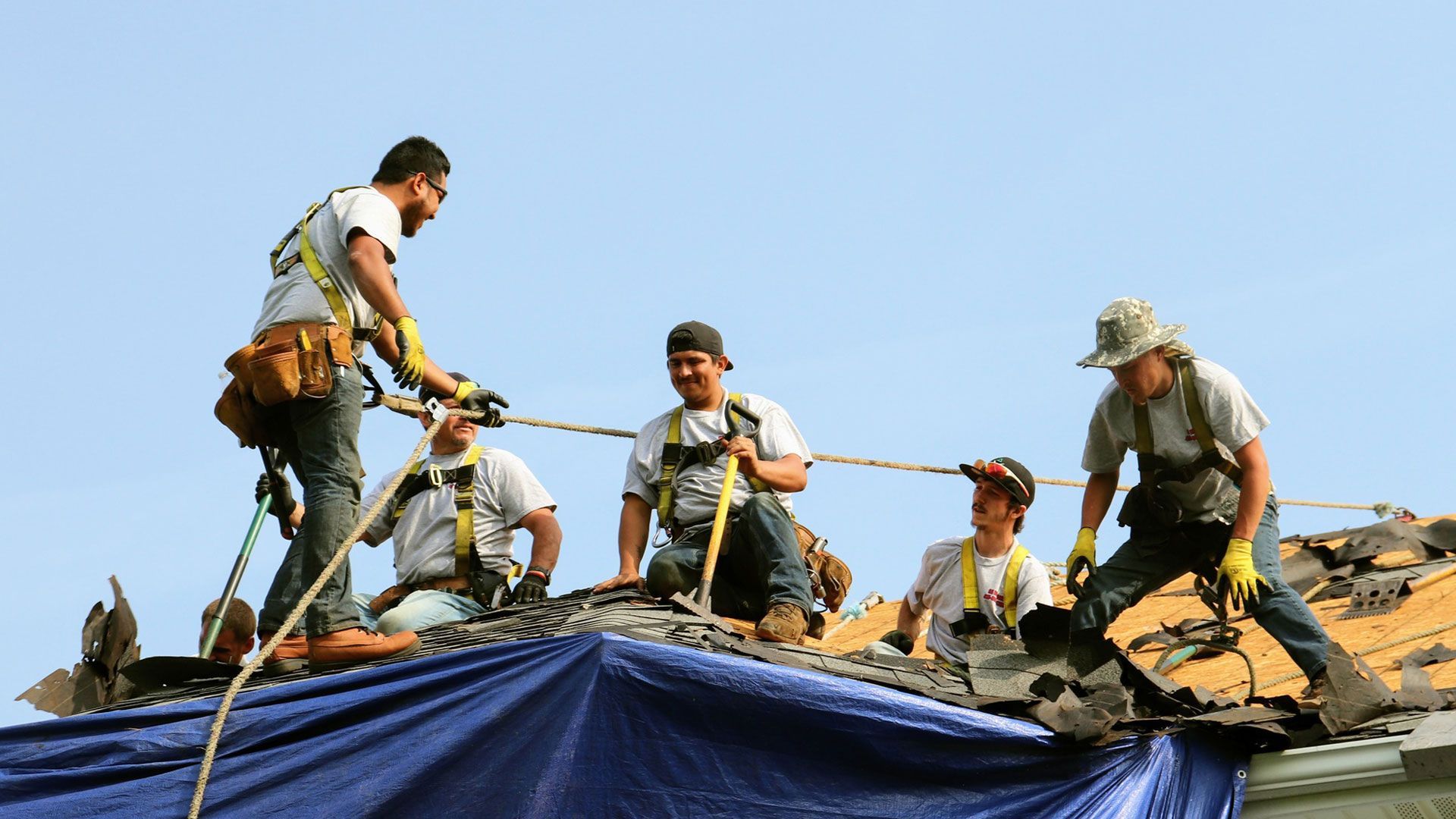 Construction workers on a roof, secured by ropes, repairing damage under a blue sky.