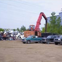 Excavator in a car scrapyard with stacked junk and parked cars on a dirt lot