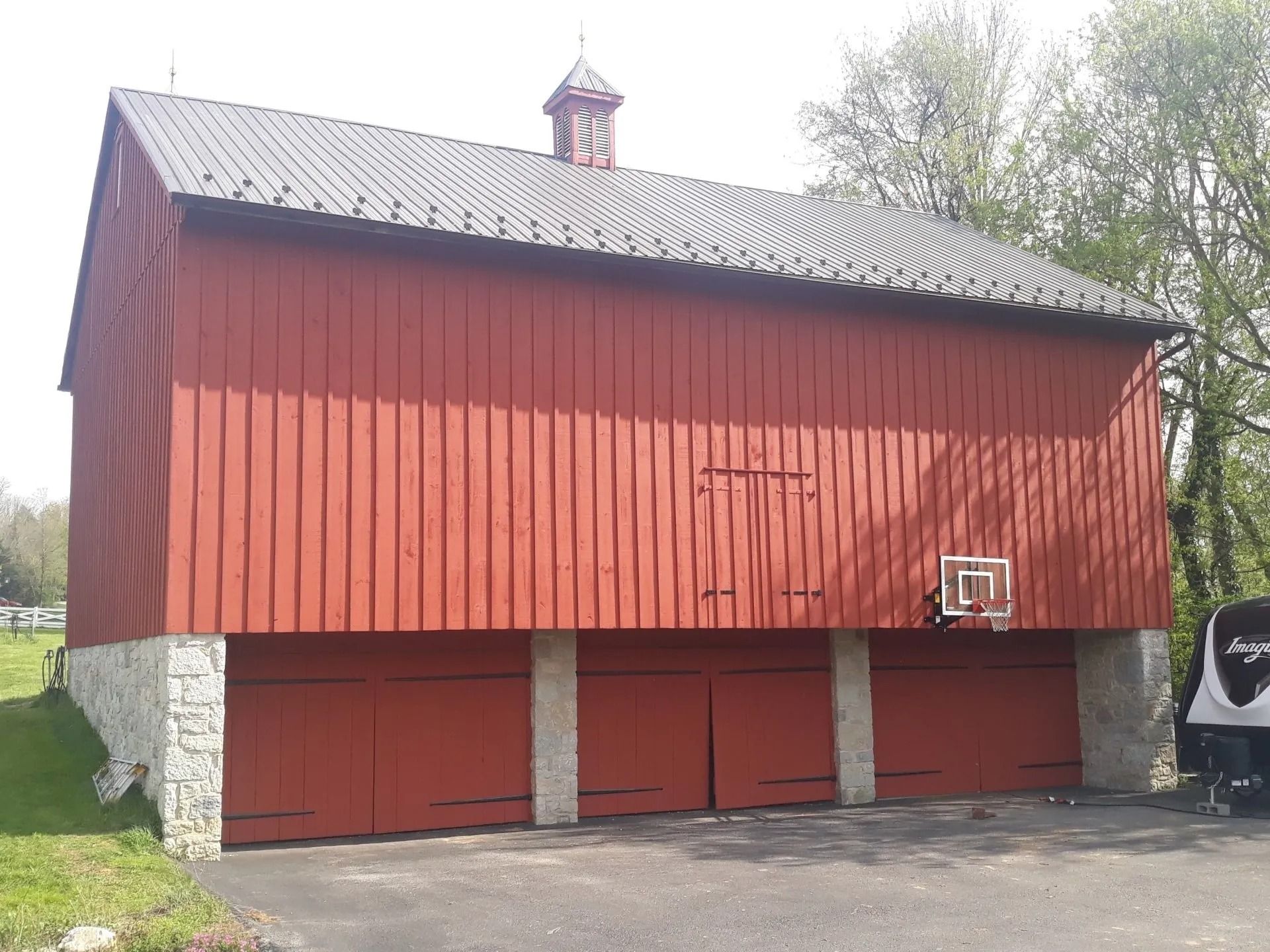 Red barn with gray roof, stone foundation, and three red garage doors. Basketball hoop mounted on side.