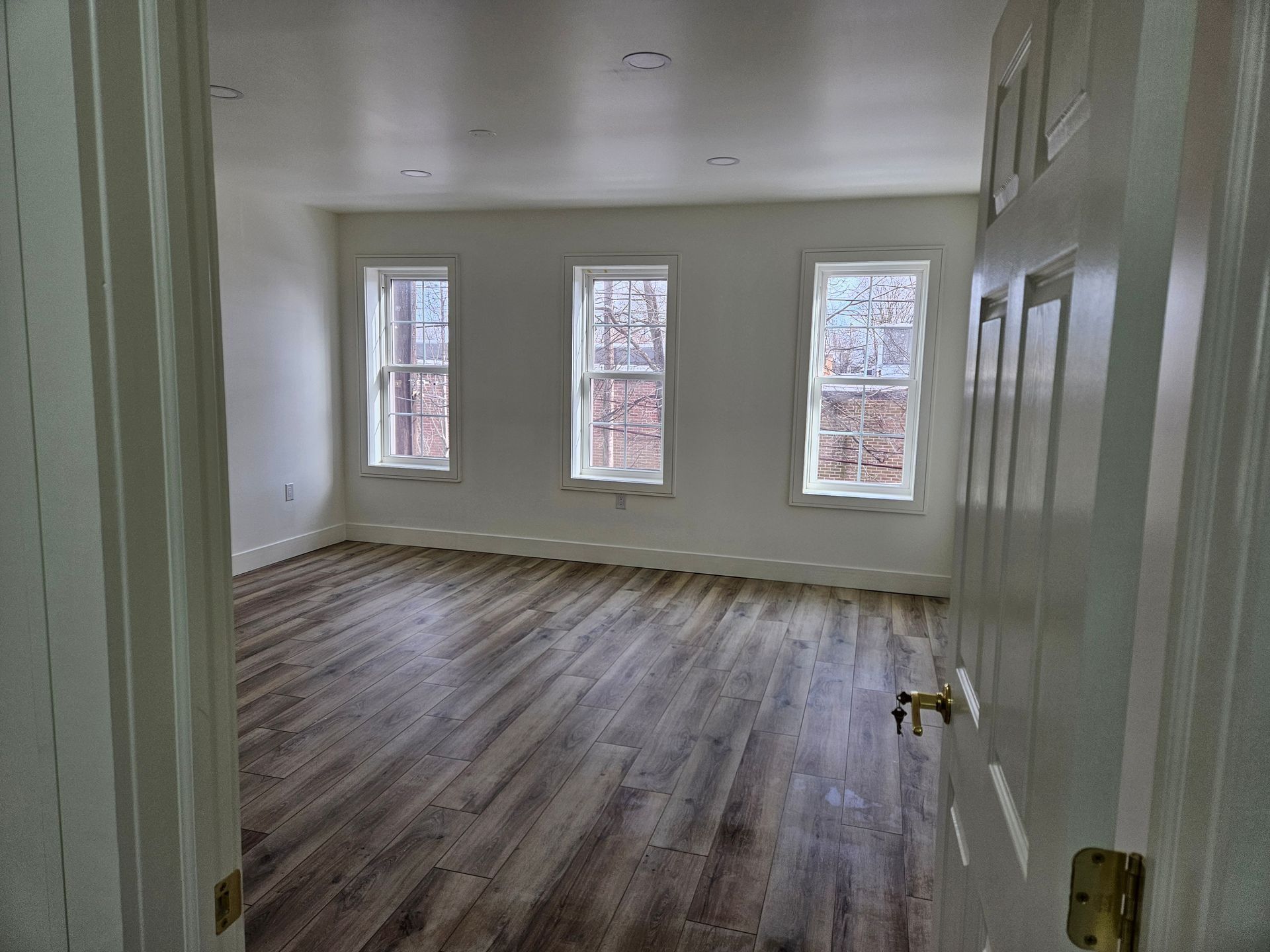 Empty, bright room with three windows, hardwood floors, and open door.