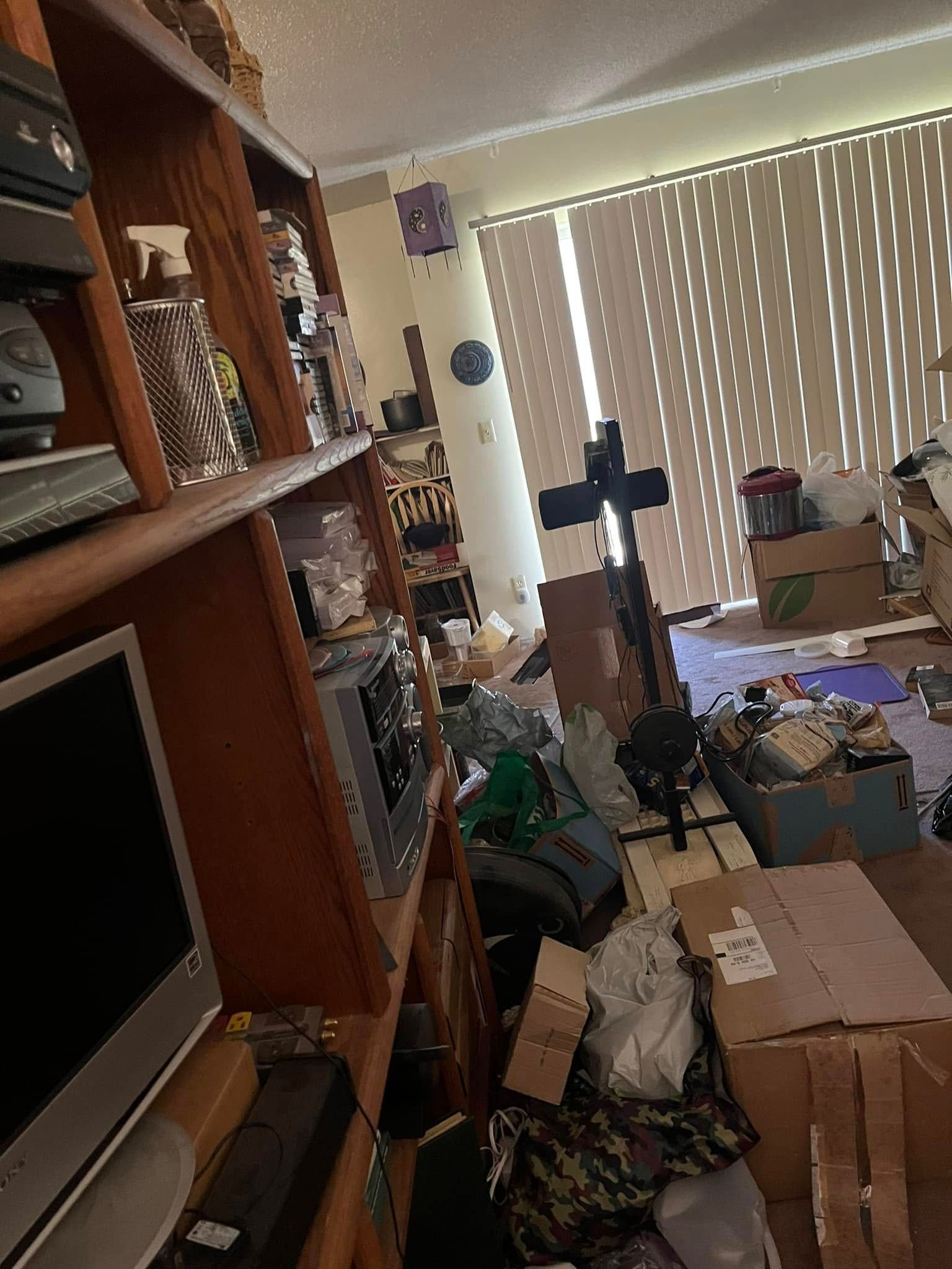 Cluttered room with a bookshelf on the left. Boxes and miscellaneous items litter the floor, and blinds cover the window.