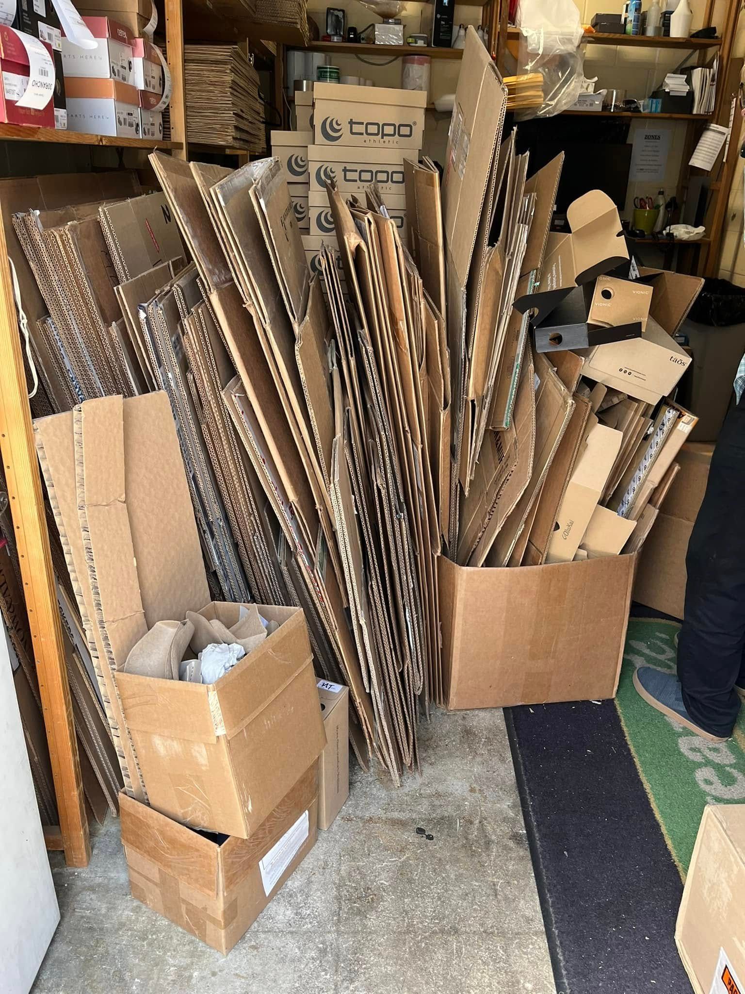 Cardboard boxes and pieces of cardboard piled inside a cluttered space, possibly a storage area or workshop.