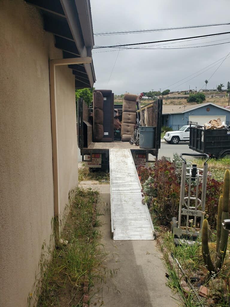 A flatbed truck loaded with furniture is parked on a driveway next to a house. A ramp extends from the truck to the ground.
