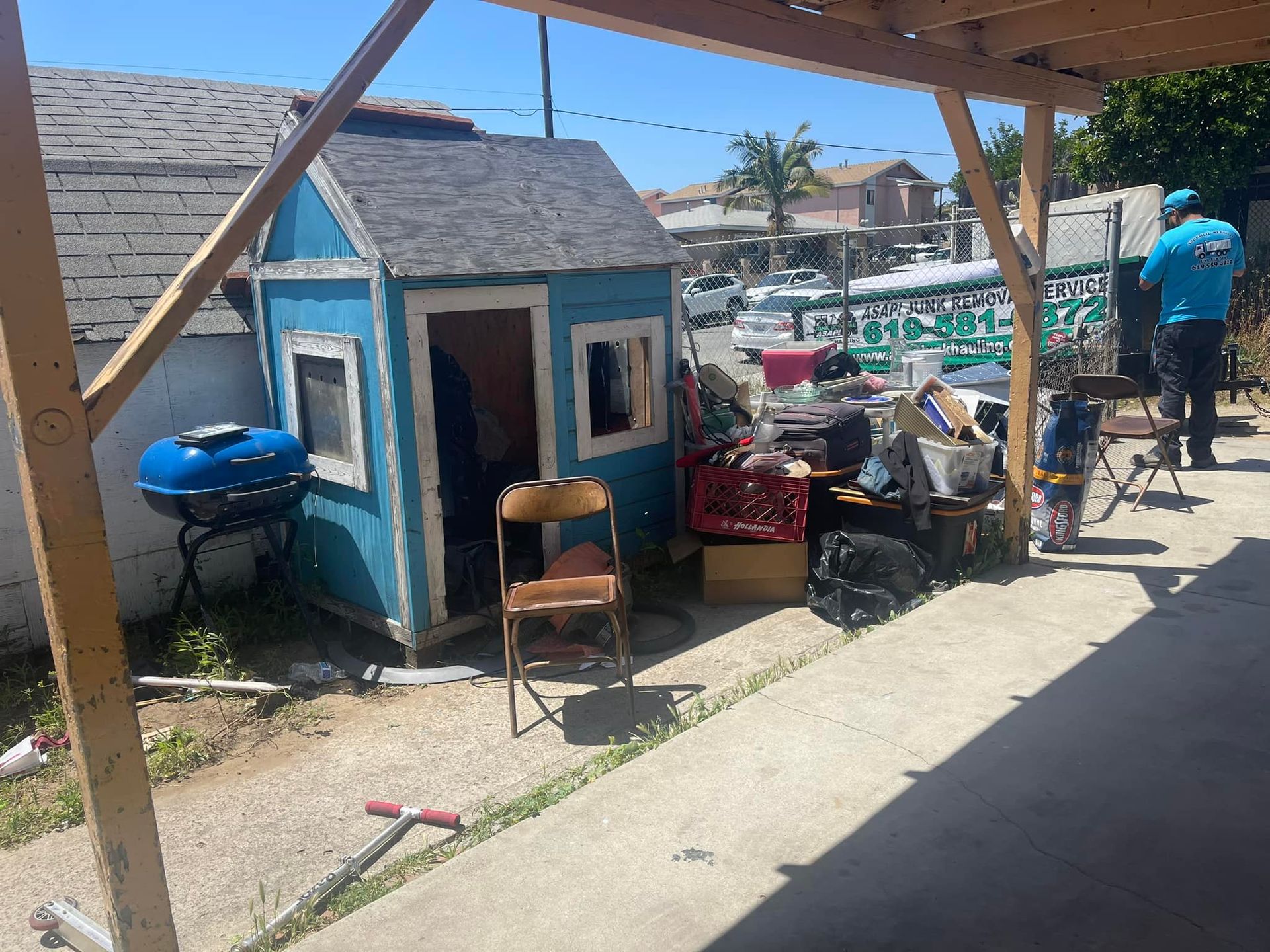Backyard with a blue playhouse, assorted items, and a person working. A small child is also present.