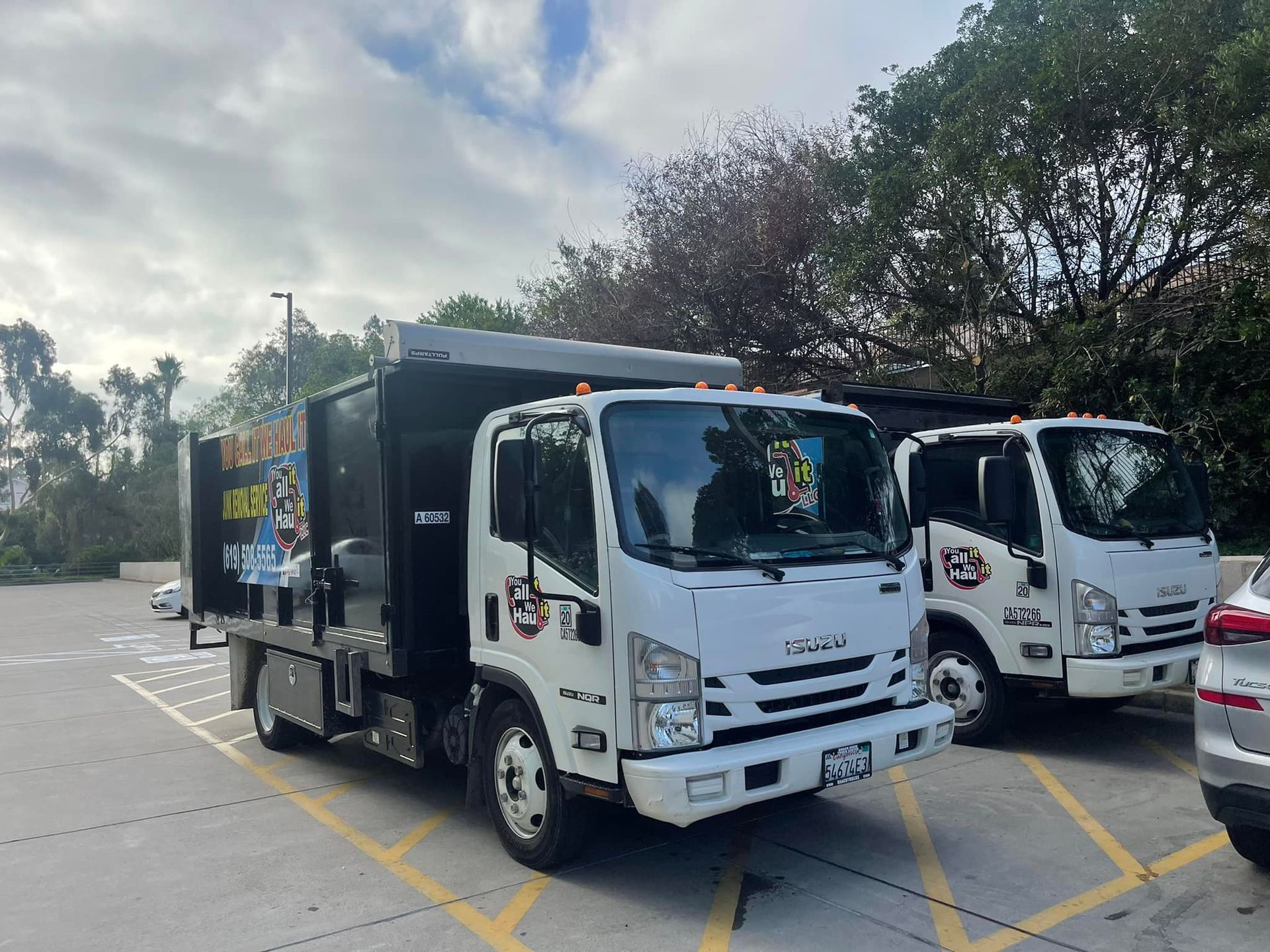 Two white work trucks parked in a parking lot; one is black-boxed in the back. Trees and a cloudy sky are in the background.