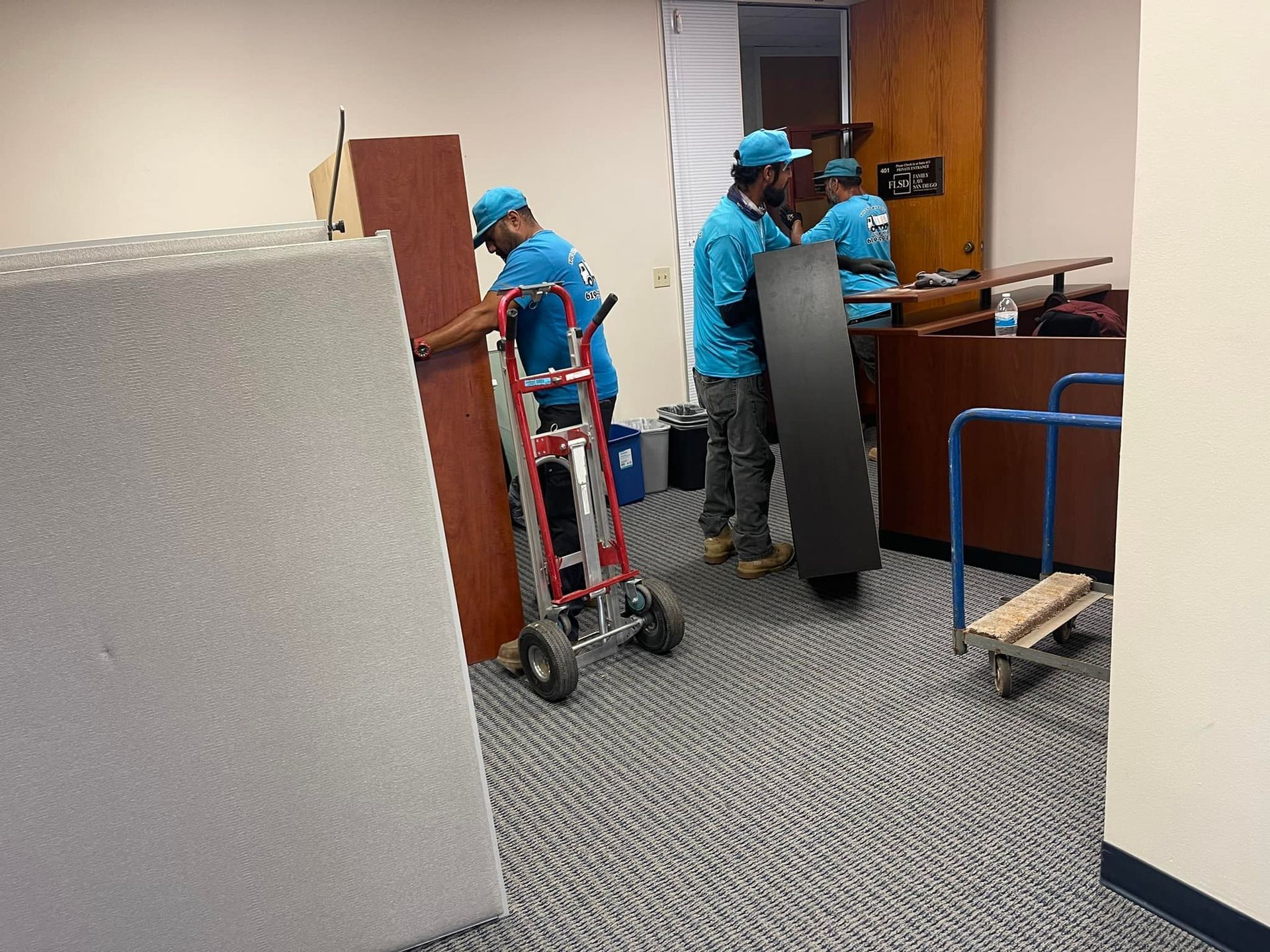 Three movers in blue shirts and caps are carrying furniture in an office. One uses a dolly.
