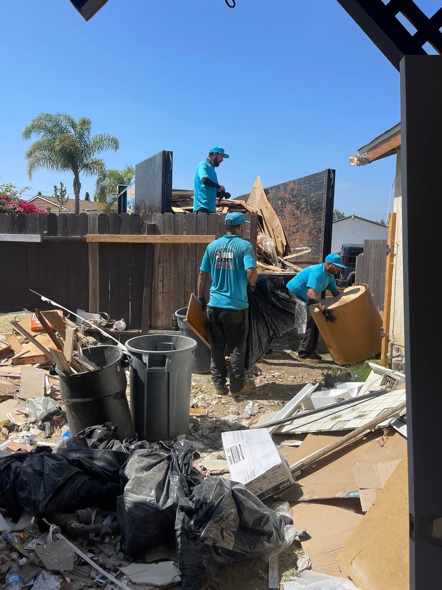 Workers in blue shirts clearing debris in a backyard under a blue sky; a fence and partial structures are visible.