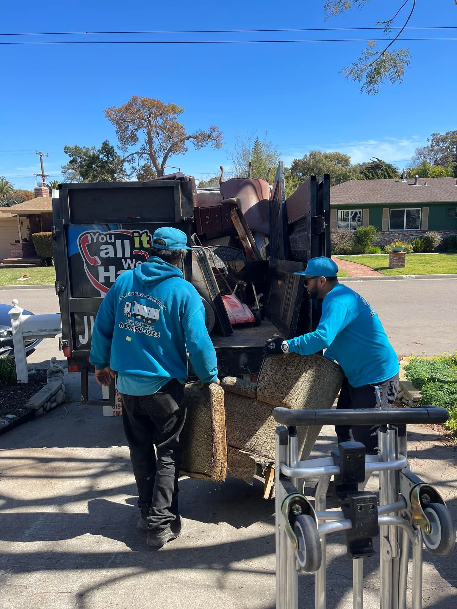 Two men in blue unloading furniture into a junk removal truck on a sunny residential street.