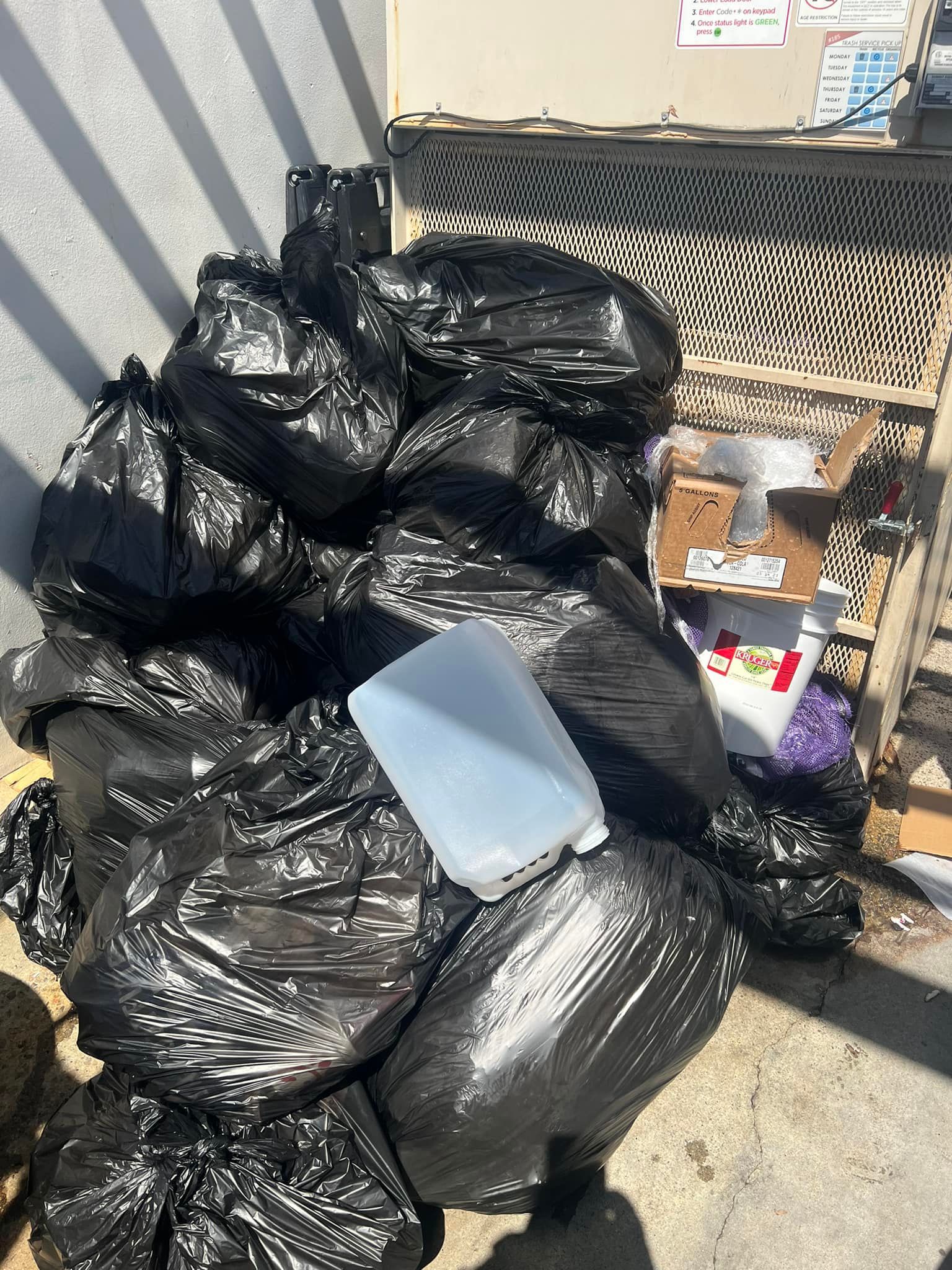Pile of black trash bags with a white water jug on top, next to a building and a metal grate.