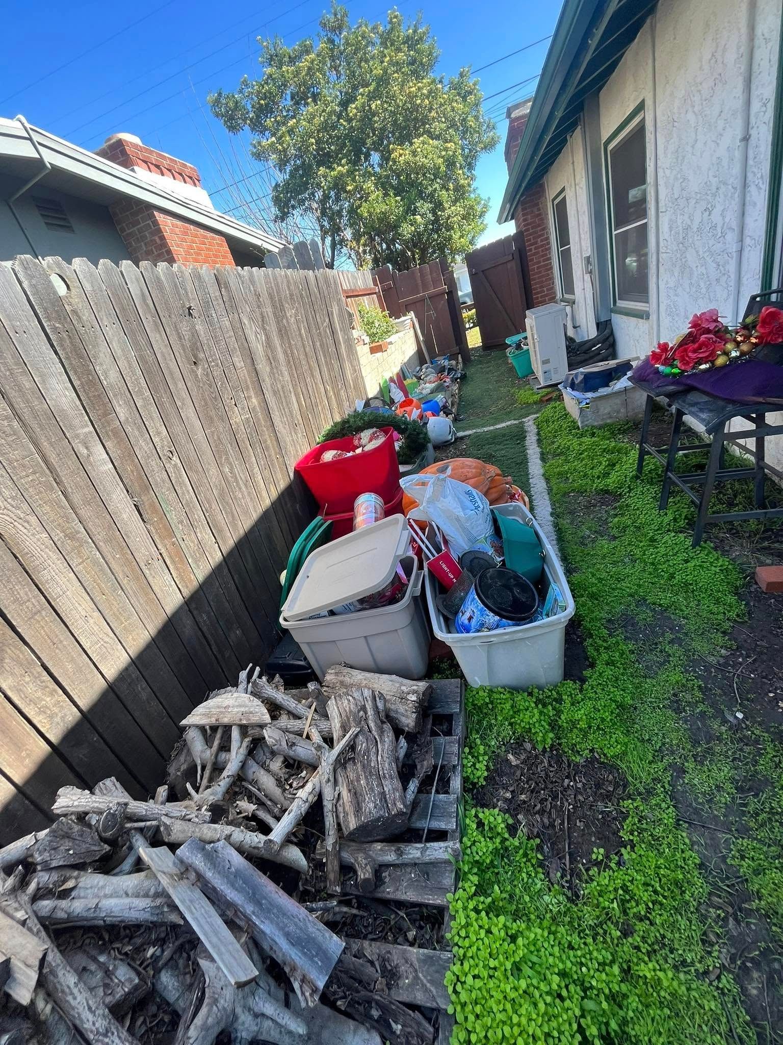 Backyard filled with clutter, including plastic bins overflowing with items and a pile of wooden debris, along a fence and side of a house.
