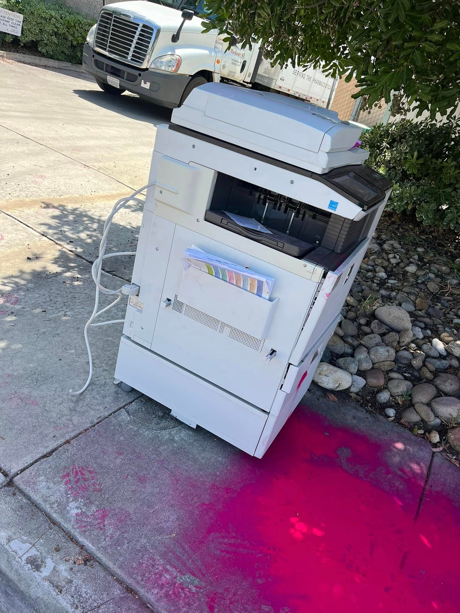 A large, white copier sits on a sidewalk, with spilled pink toner covering the pavement. A truck is visible in the background.