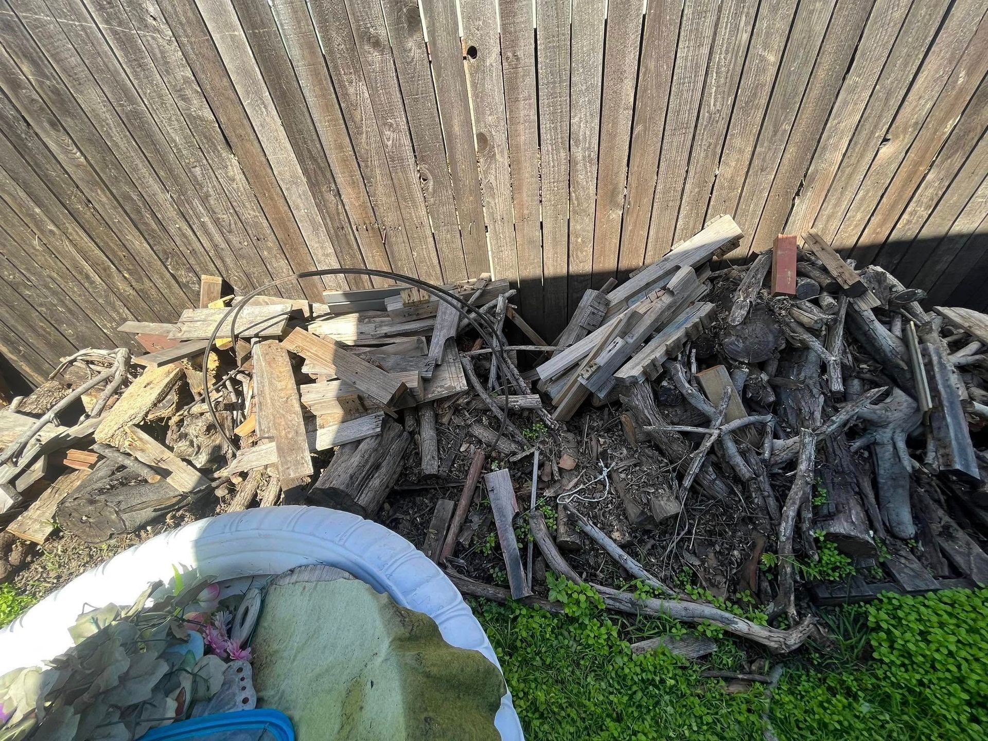 Pile of firewood and debris next to a weathered wooden fence, with some greenery in the foreground.