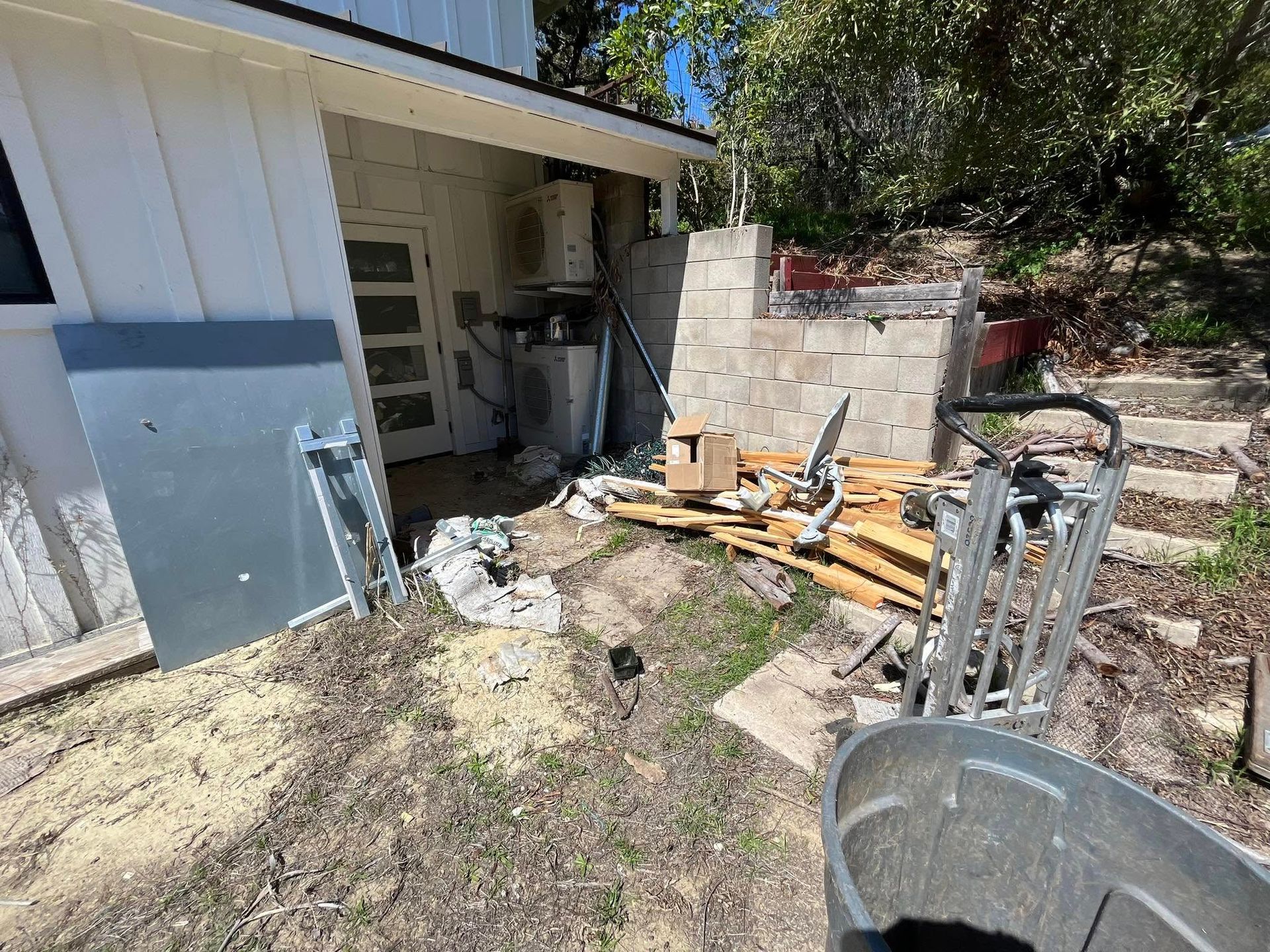 A cluttered outdoor area with construction debris near a white building and a stone structure. A gray metal sheet leans against the building.