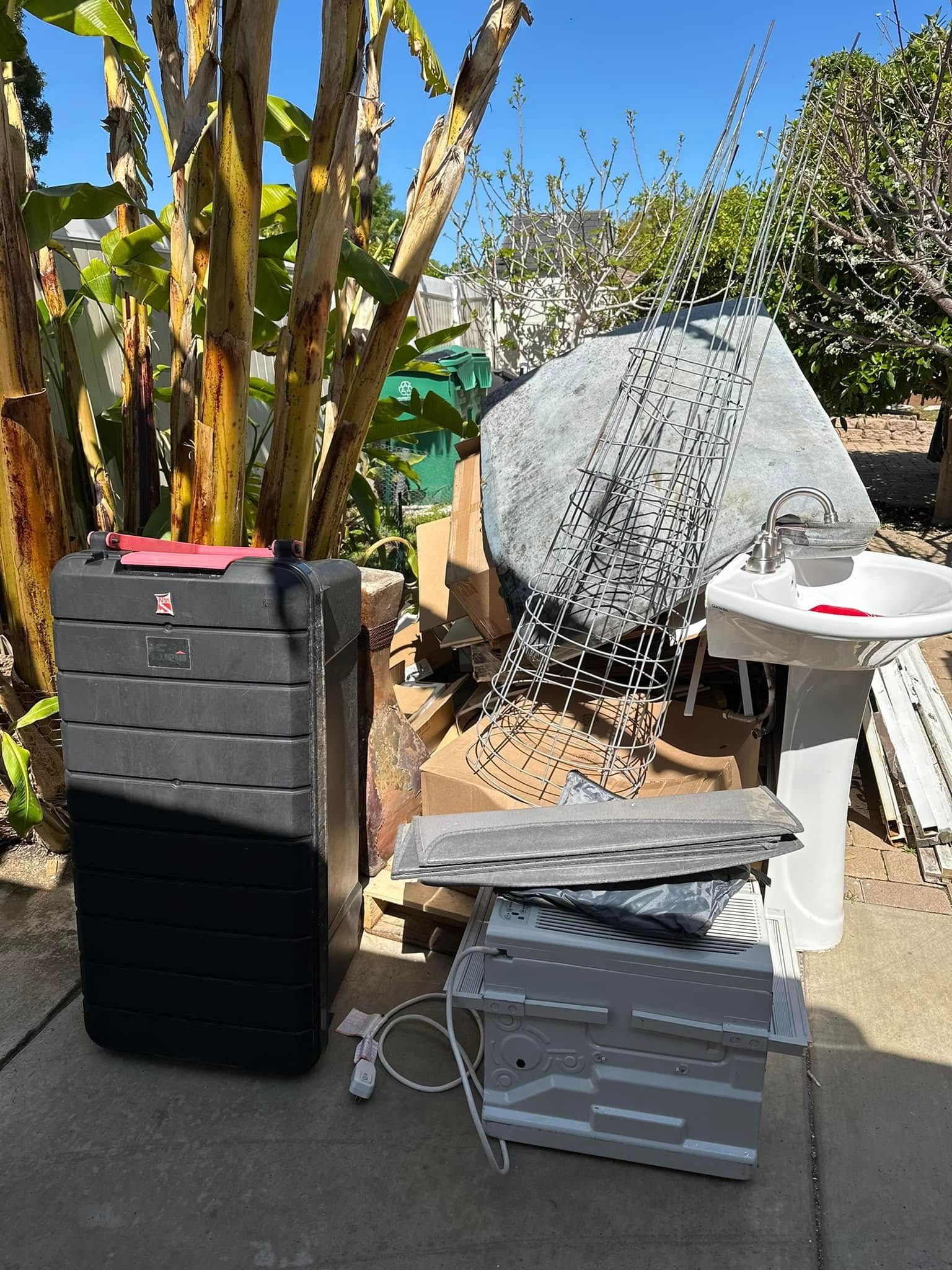 A black compost bin and pile of construction debris in a sunny yard, including a sink and large rock.