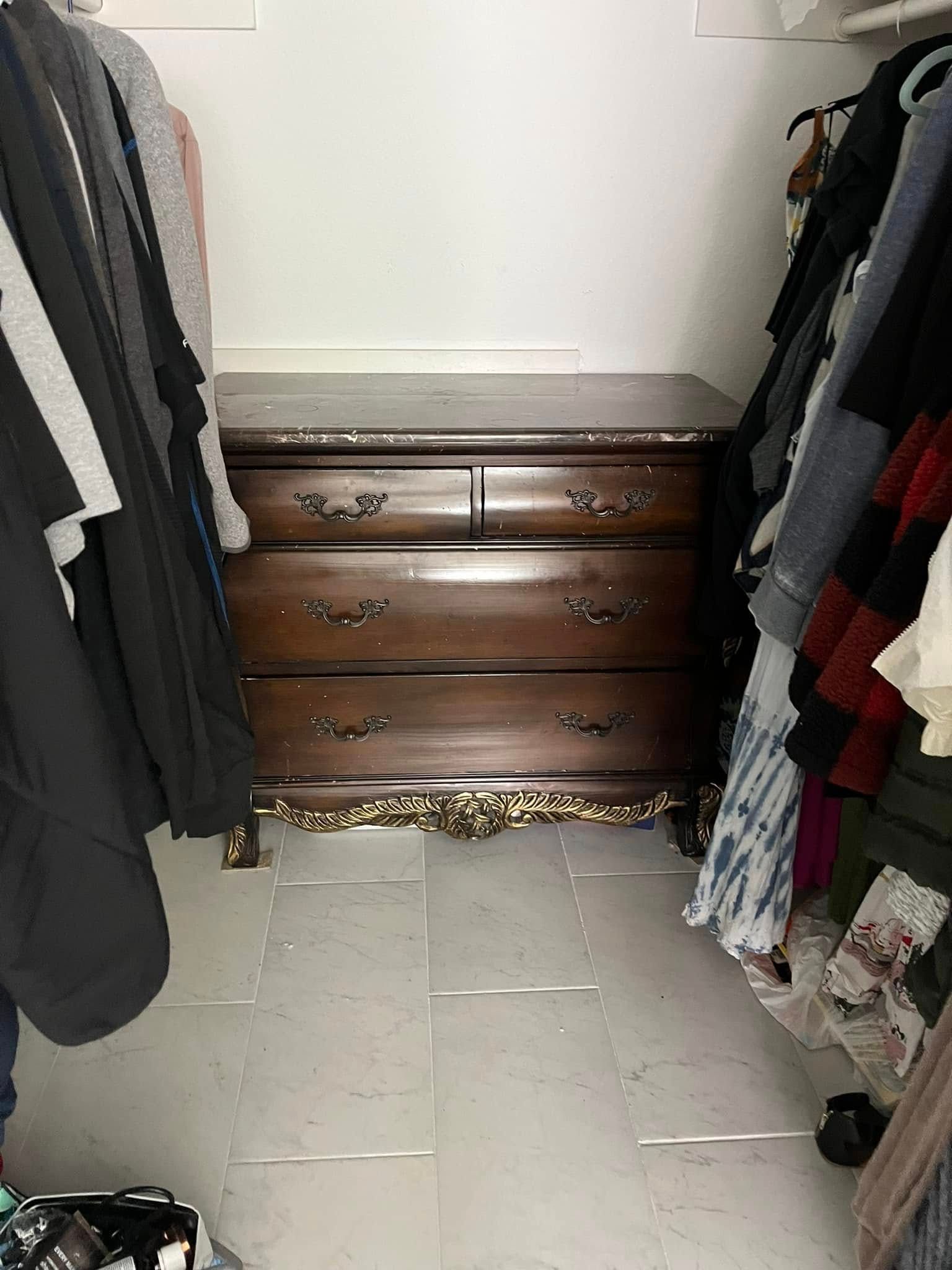 A dark wood dresser with four drawers sits between two rows of hanging clothes in a closet.