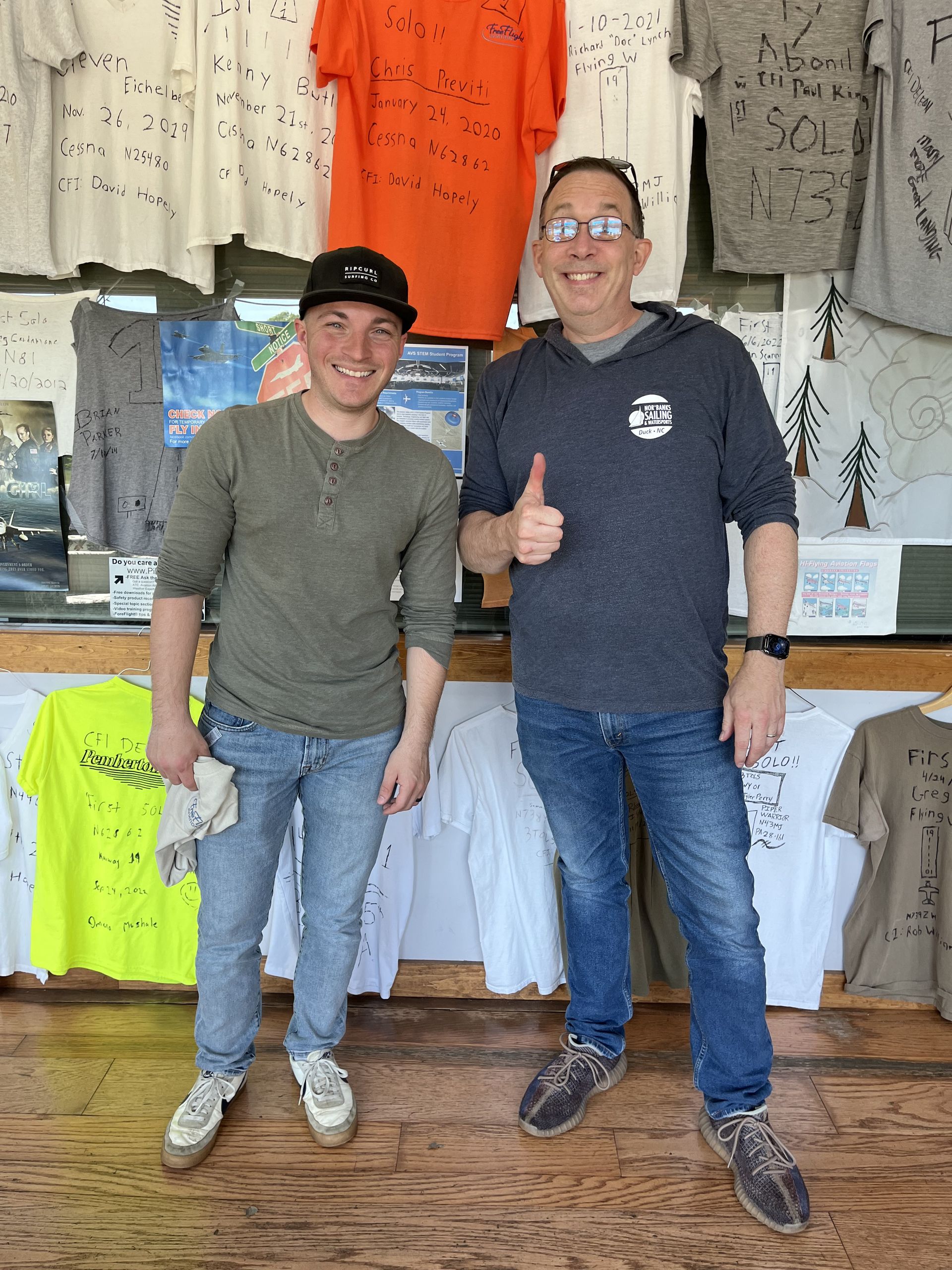 two men are standing in front of a display of shirts and one is giving a thumbs up