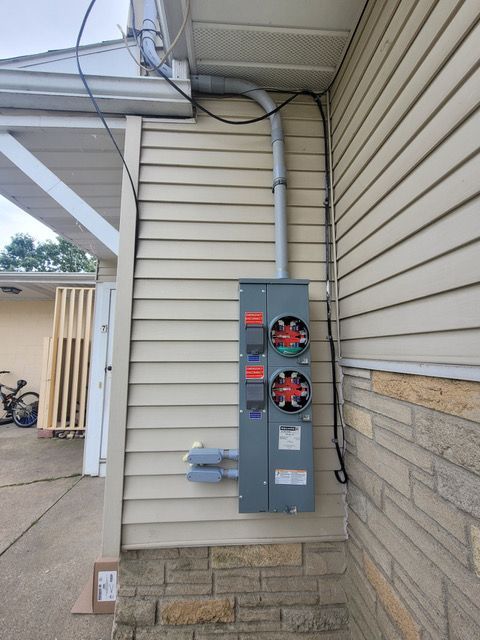 Exterior electrical meter and panel on a building with beige siding and brick foundation.