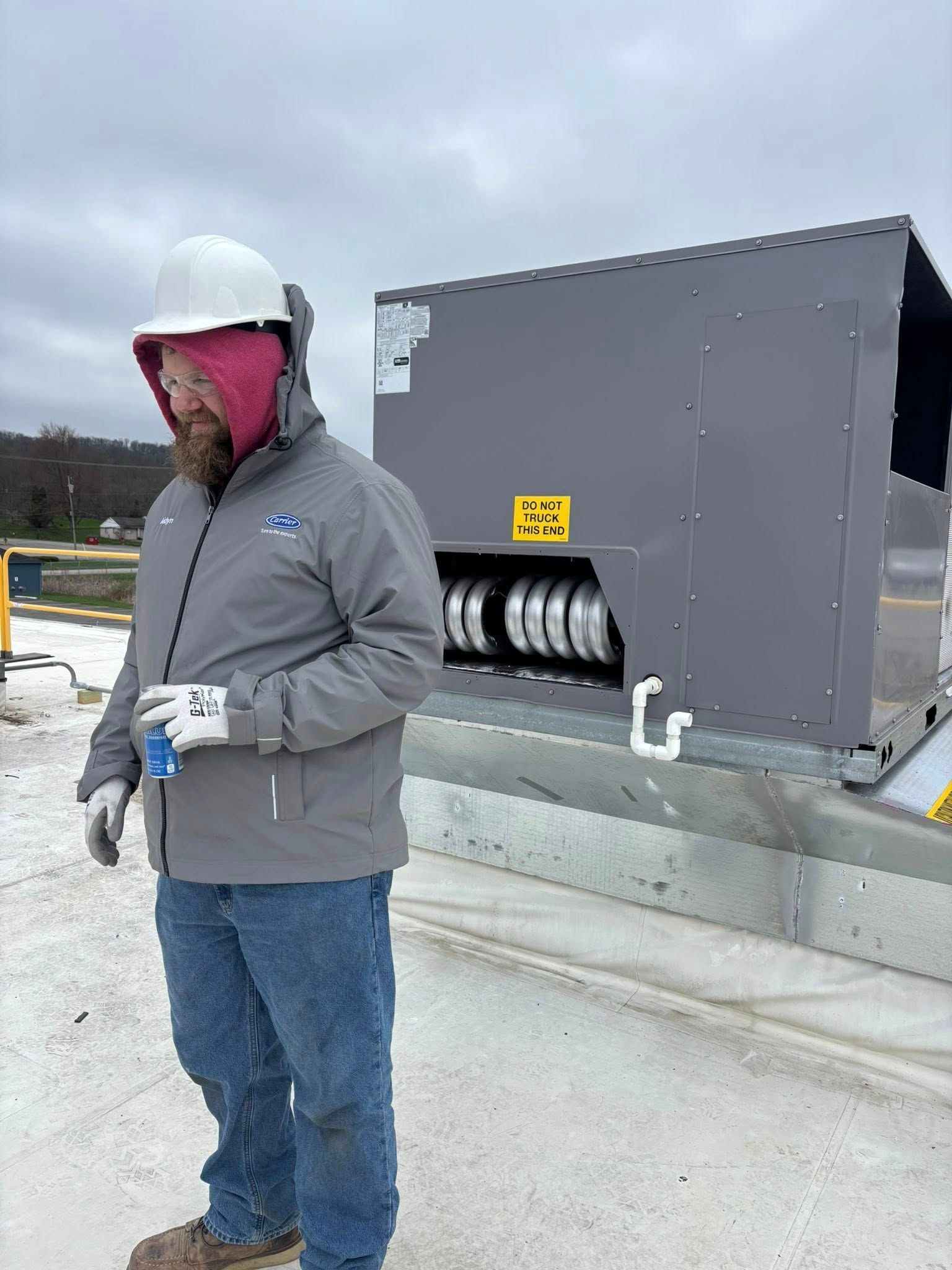 Man in a hard hat and jacket stands near HVAC equipment on a rooftop. Cloudy sky.