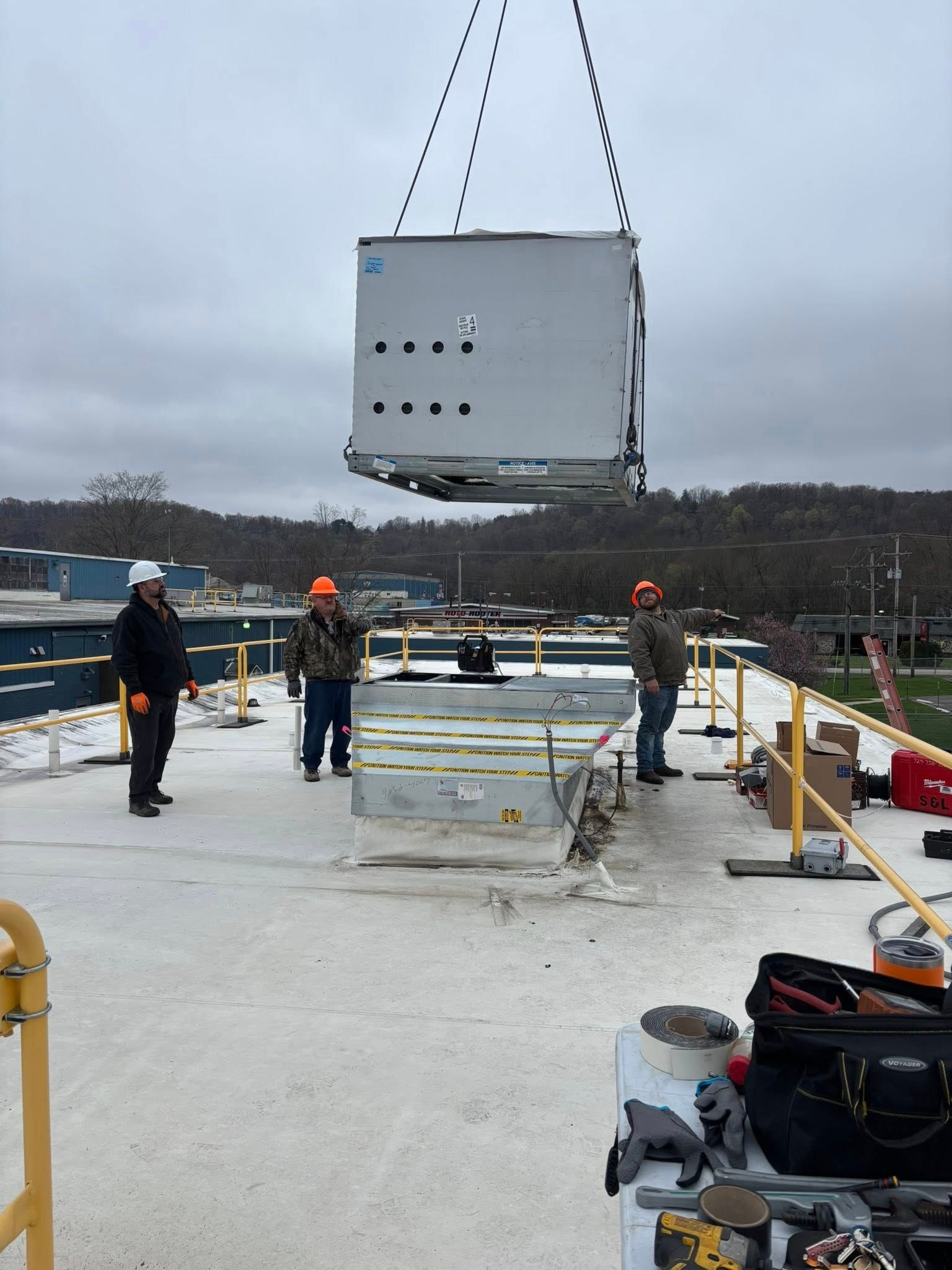 Workers on a rooftop oversee a crane lifting an HVAC unit. Cloudy sky, industrial setting.