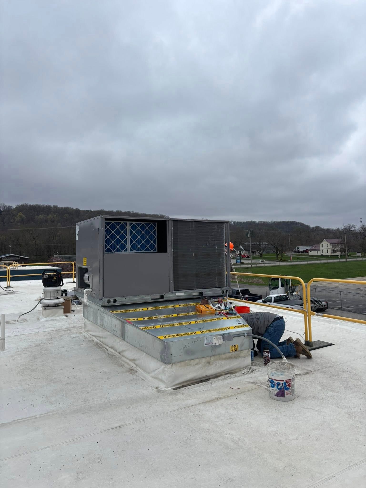HVAC unit on a flat roof, worker in blue overalls, cloudy day.