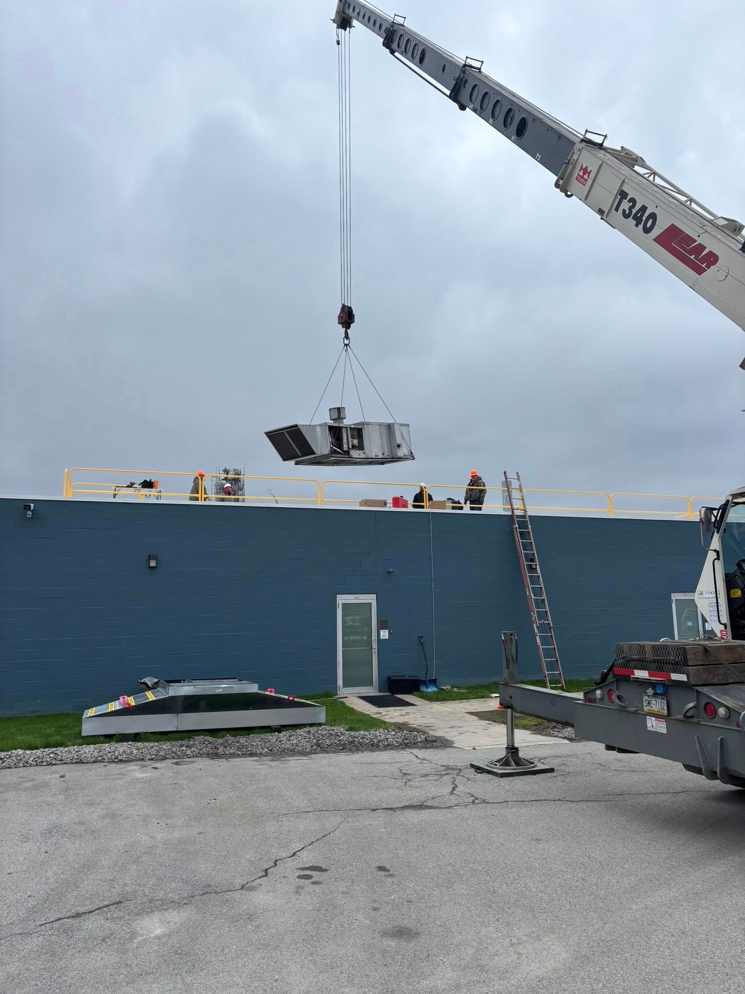 Crane lifting an HVAC unit onto a building's roof with workers present. Gray sky, blue building.