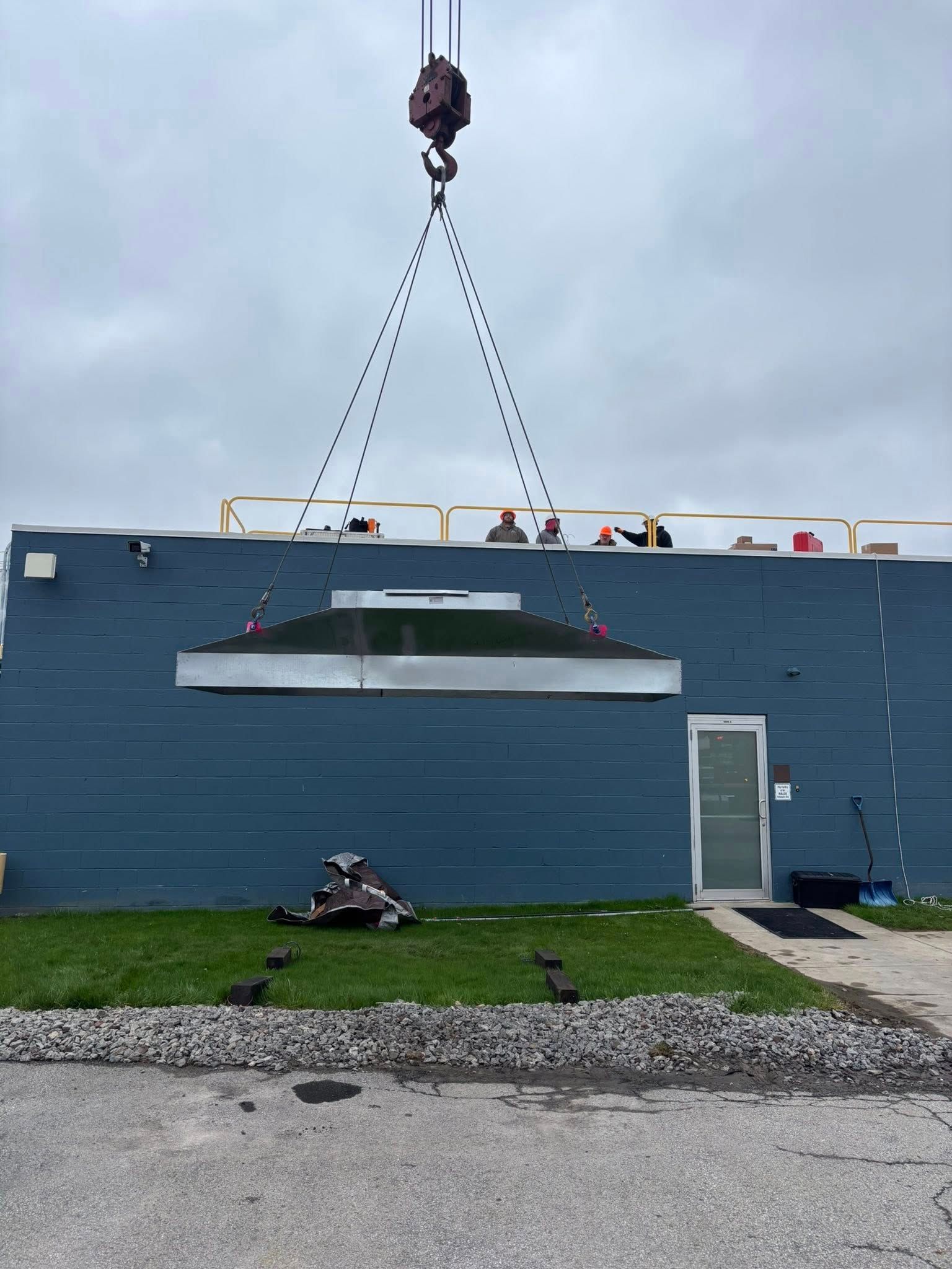 A crane lifting a large, silver roof section onto a blue building on a cloudy day.