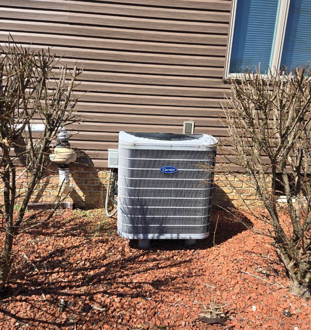 Air conditioning unit next to a house with brown siding, flanked by leafless bushes.