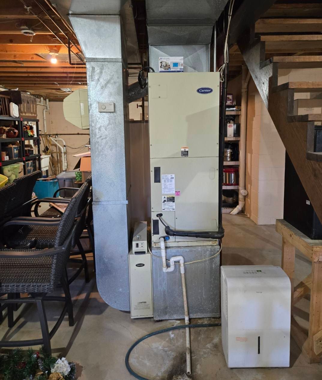 Furnace and ductwork in a basement setting. White furnace, silver ducts, wooden stairs, and a dehumidifier.