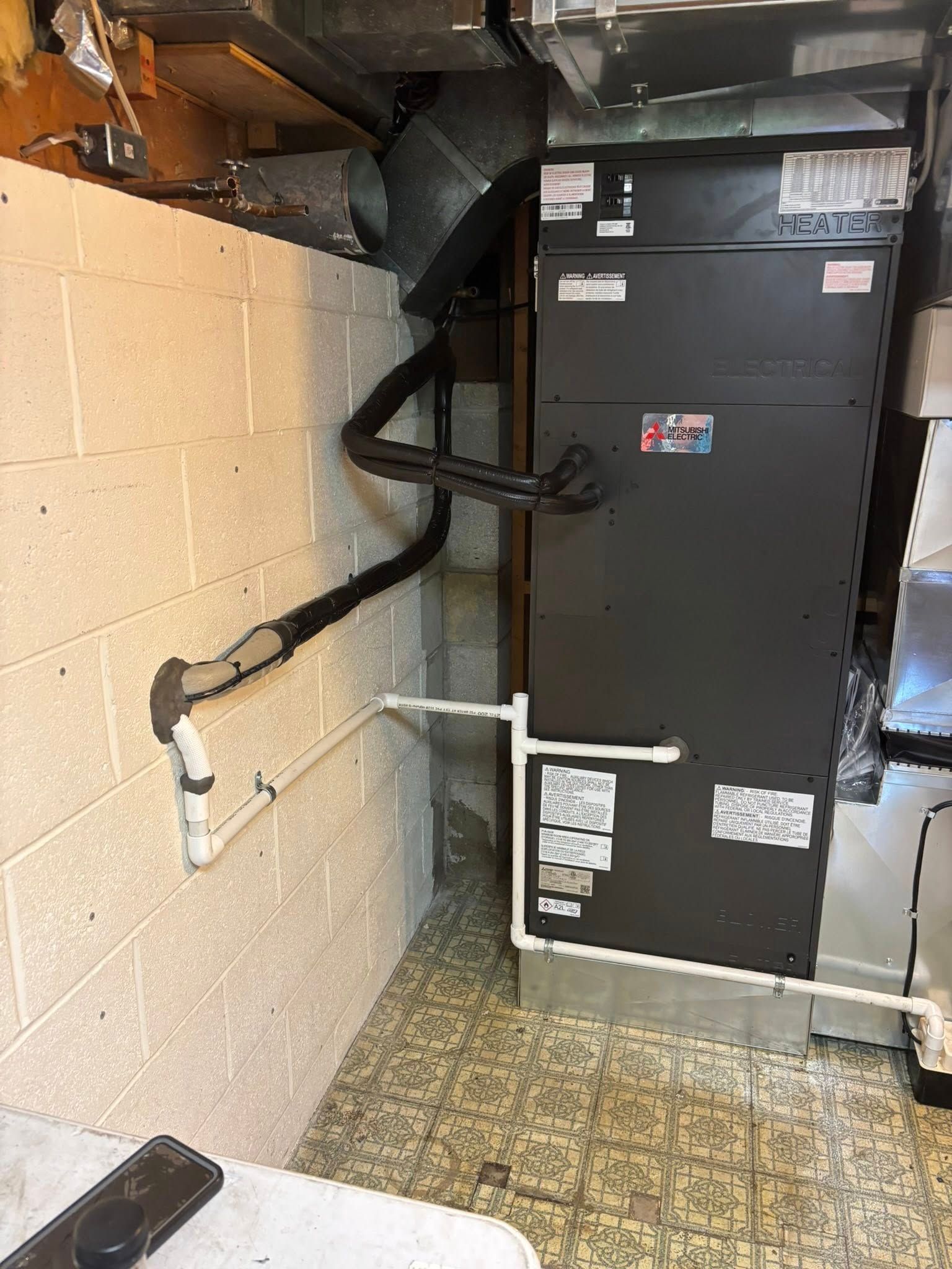 Furnace in a basement, with attached pipes and ductwork. Grey metal unit, white brick wall.