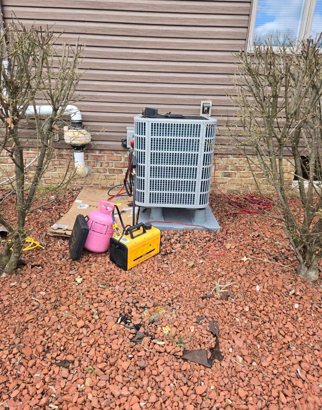 HVAC unit with tools, bushes, and a pink refrigerant tank on red gravel.