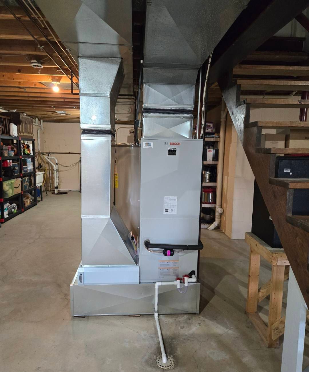 Furnace with silver ductwork in a basement. White PVC pipe extends to the floor. Wooden stairs on right.