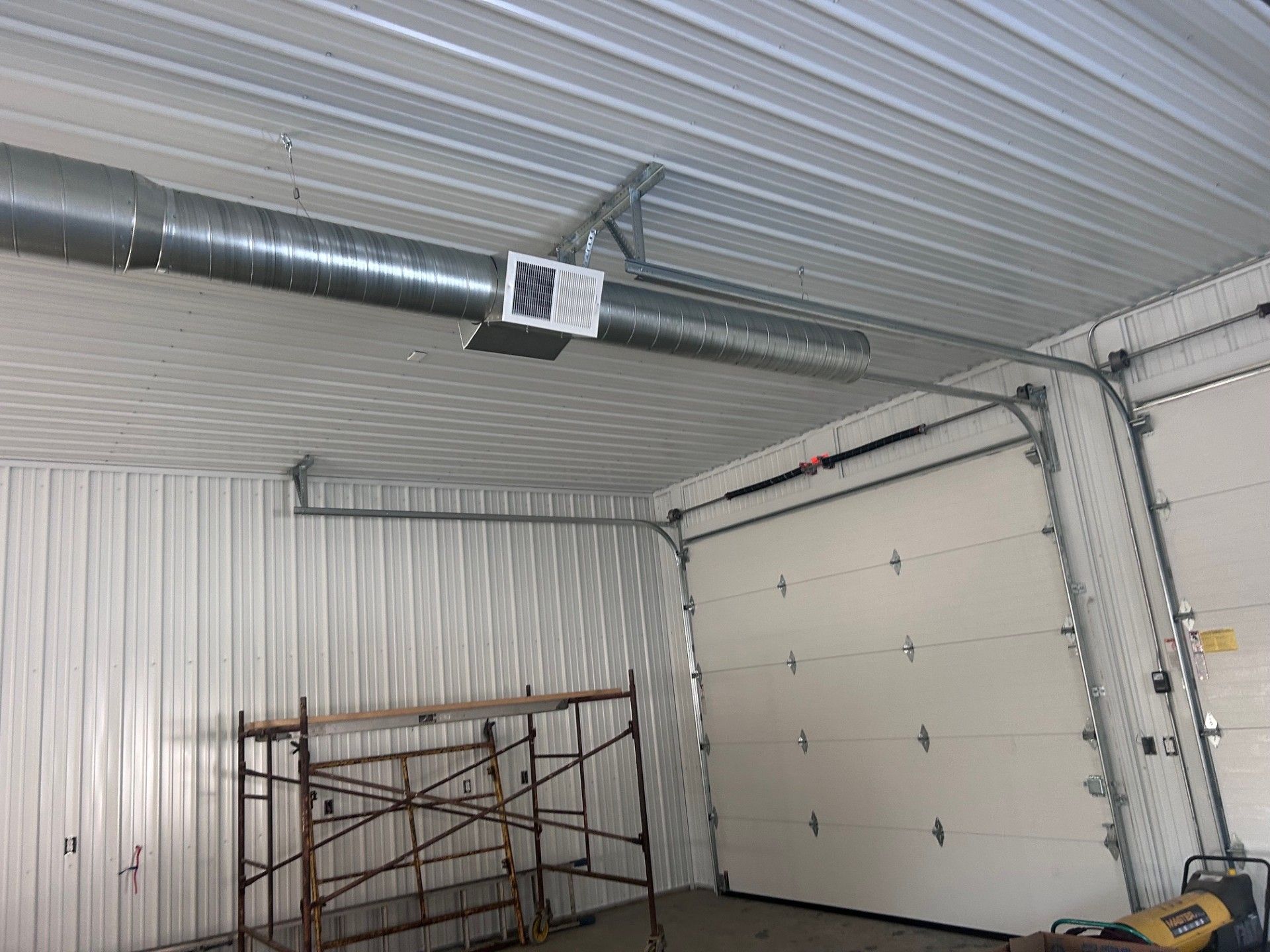 Interior of a garage with a metal roof, ductwork, a heater, and a closed garage door. Scaffolding is on the floor.