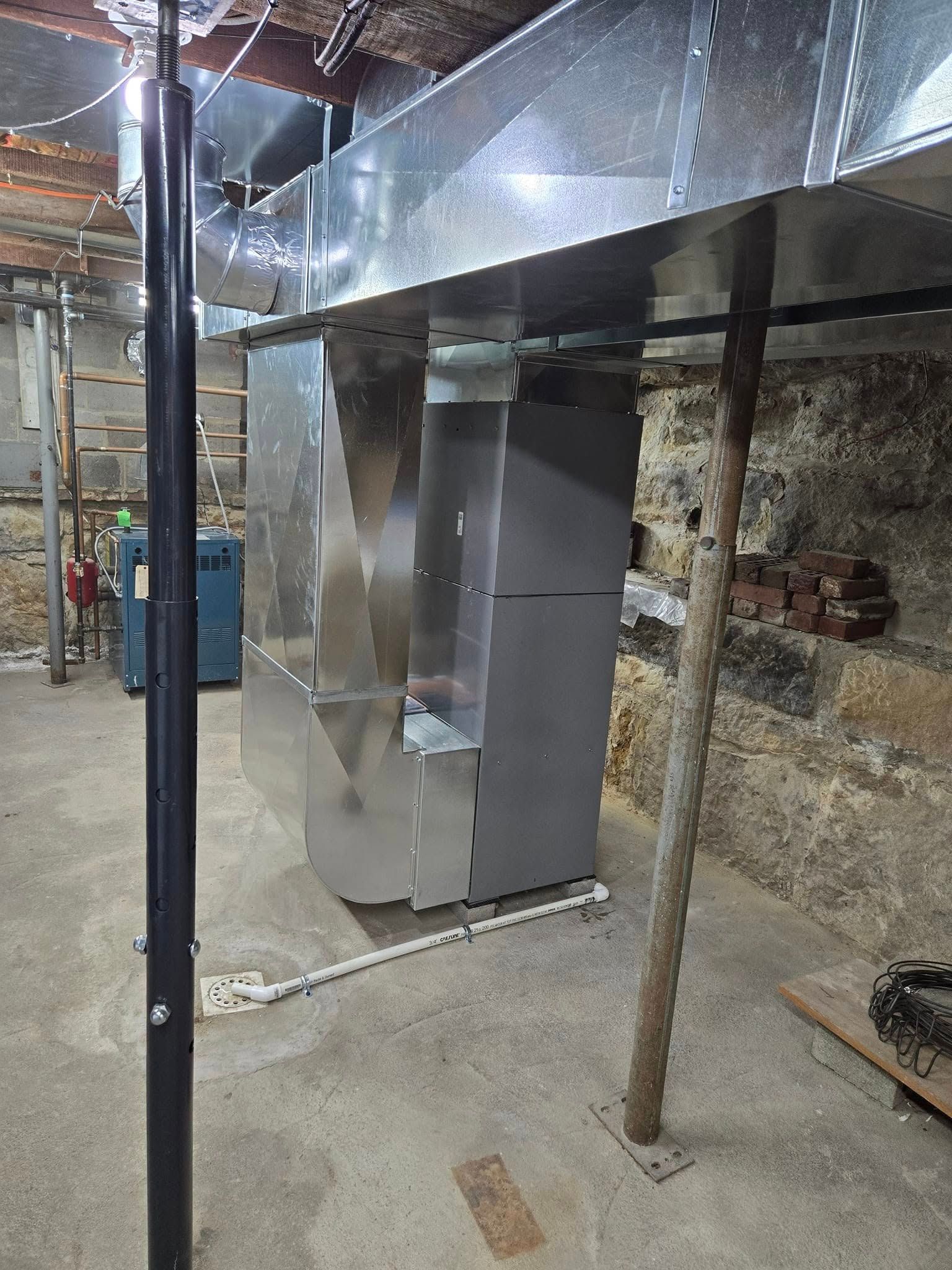 HVAC ductwork and furnace in a basement setting. Metal components, support beams, and exposed brick wall.